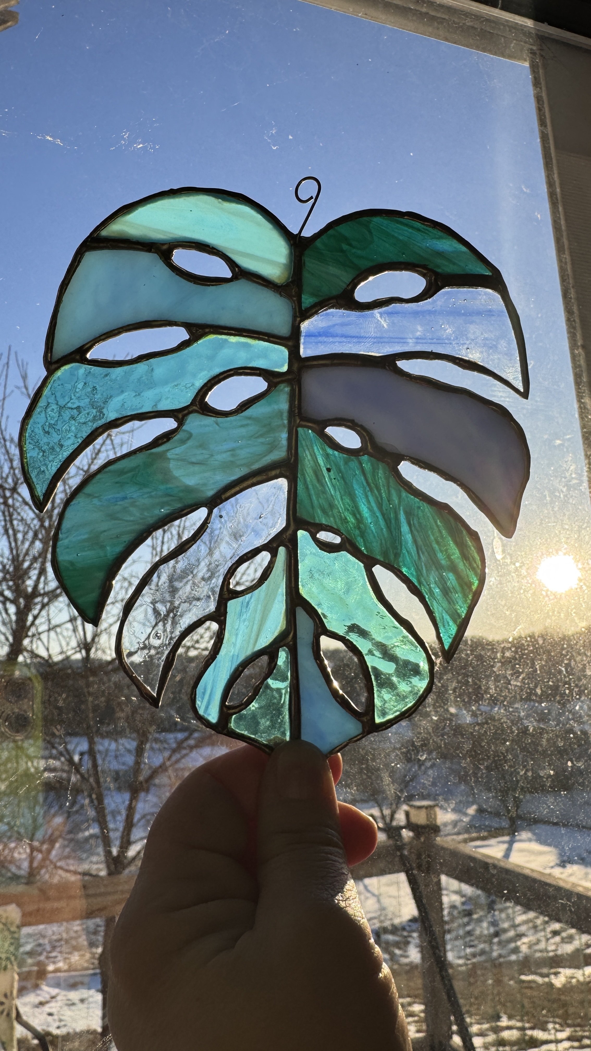 A stained glass leaf ornament held up to the sunlight, with a winter landscape seen through the window behind it.