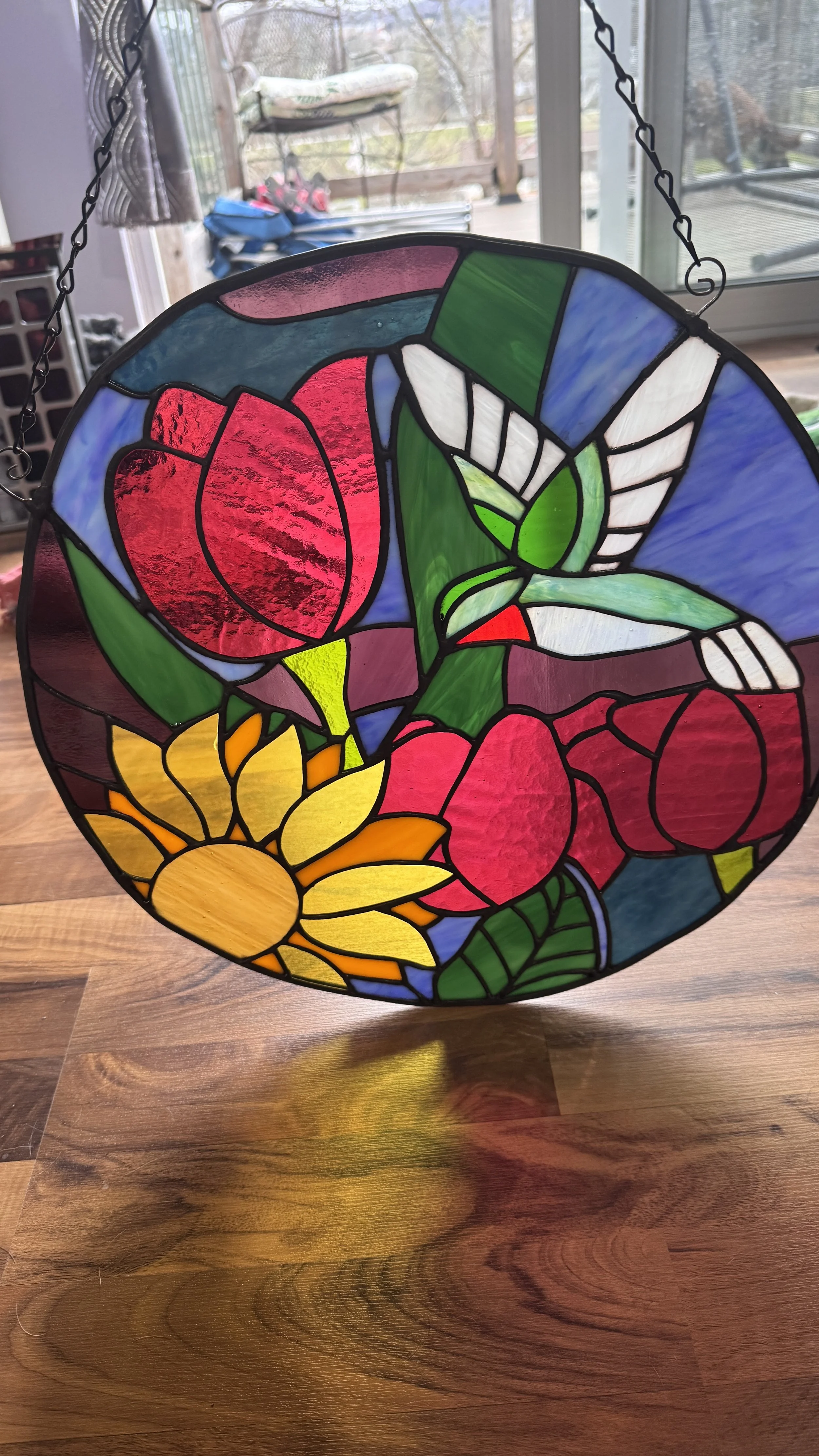 Colorful stained glass hanging with a design of a red tulip, a white flower, yellow sunflower, and green leaves, hung indoors near a window.