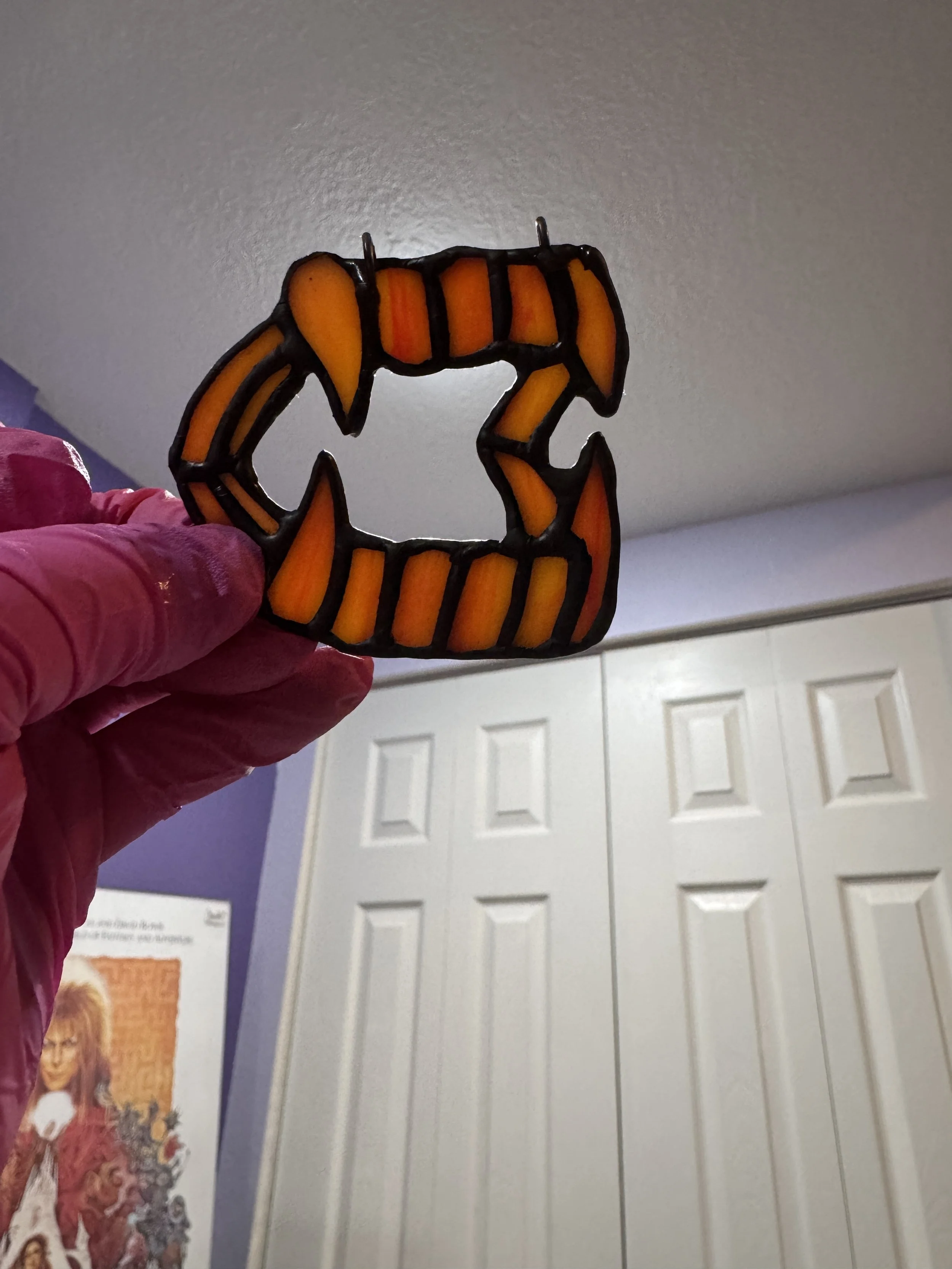 A stained glass Halloween decoration of vampire teeth being held by a person wearing a pink glove, with a purple wall and white closet doors in the background.