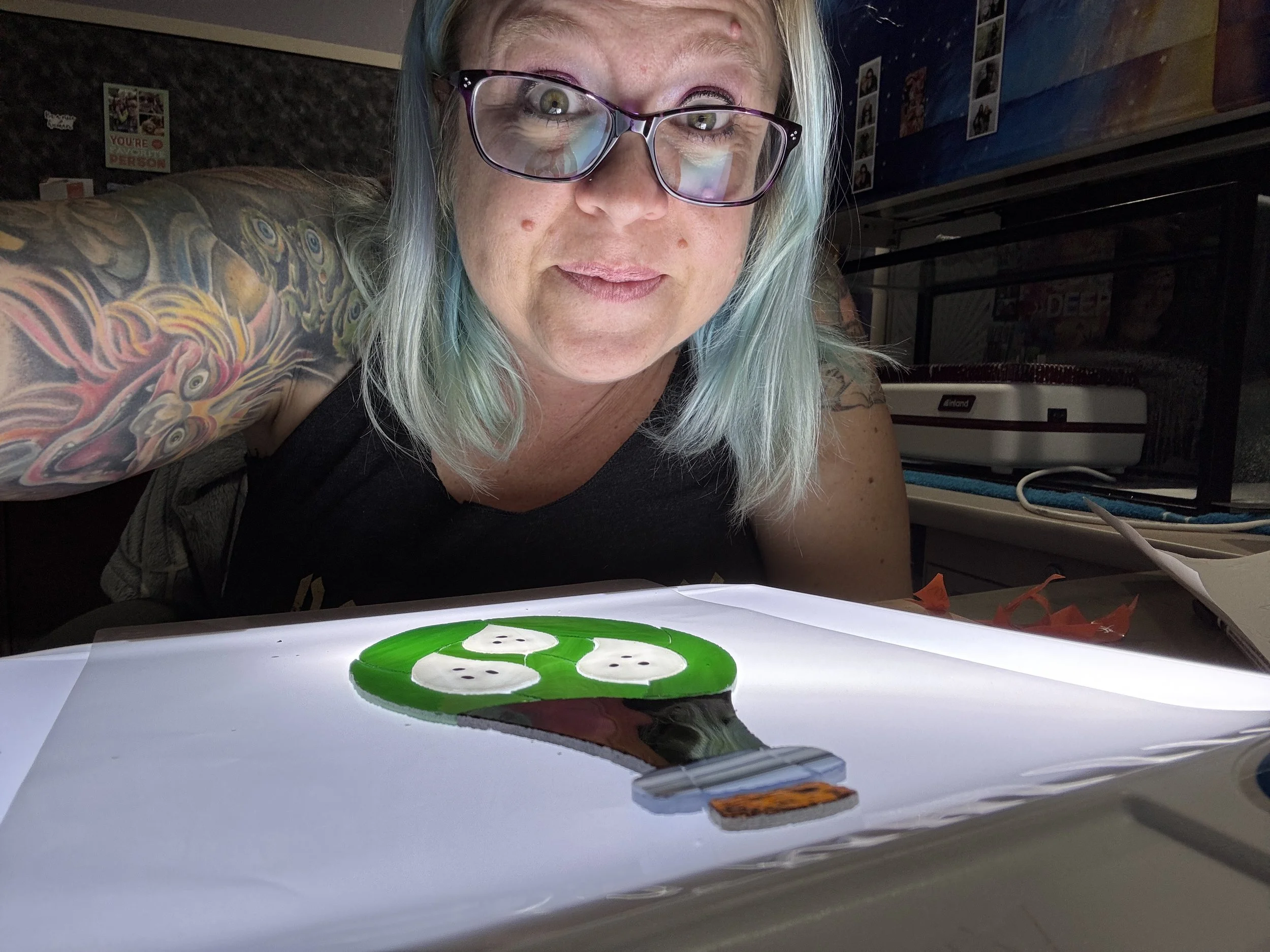 A woman with glasses and colorful hair taking a selfie behind an illuminated piece of art featuring a green cartoon character and a lighter-colored object. She has tattooed arms and is in a room with posters and various items in the background.