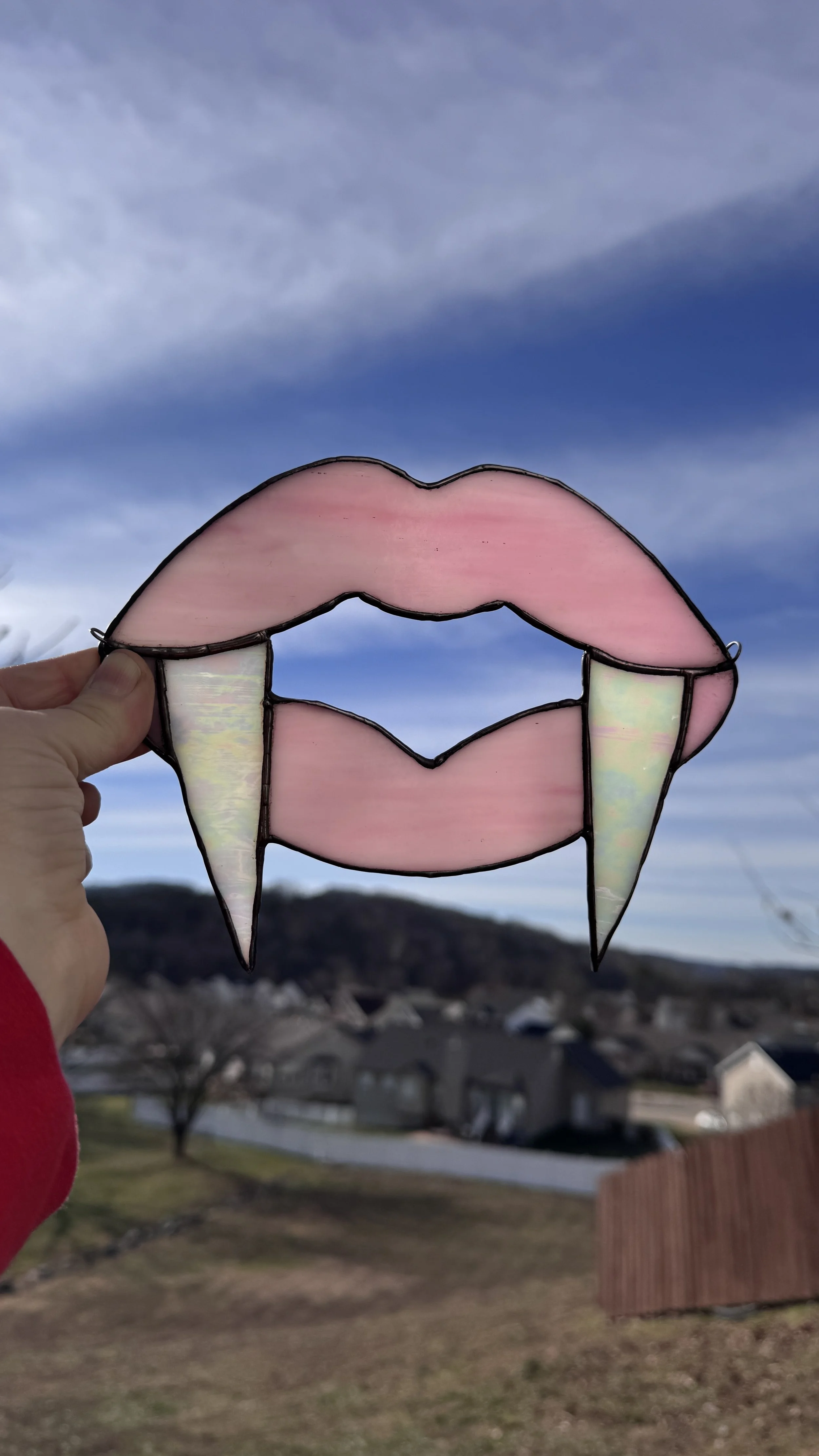A stained glass art piece shaped like lips with vampire fangs, being held up against a cloudy sky with houses and trees in the background.