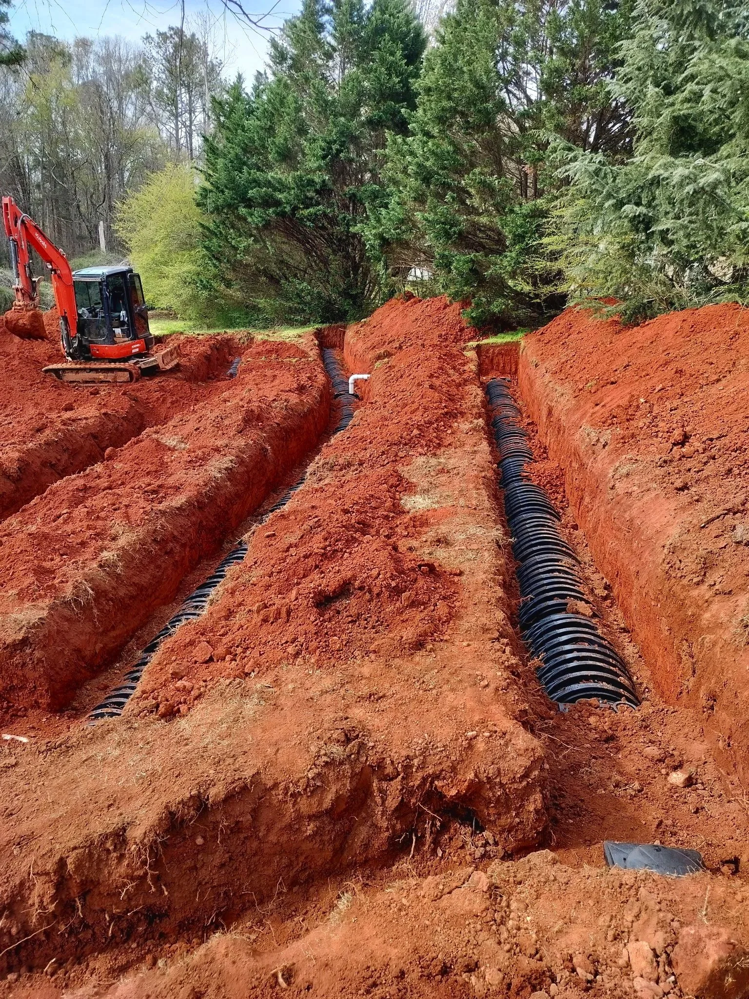 Construction site with deep trenches and black corrugated piping, a small red excavator on the left, green trees in the background, and a patch of grass.