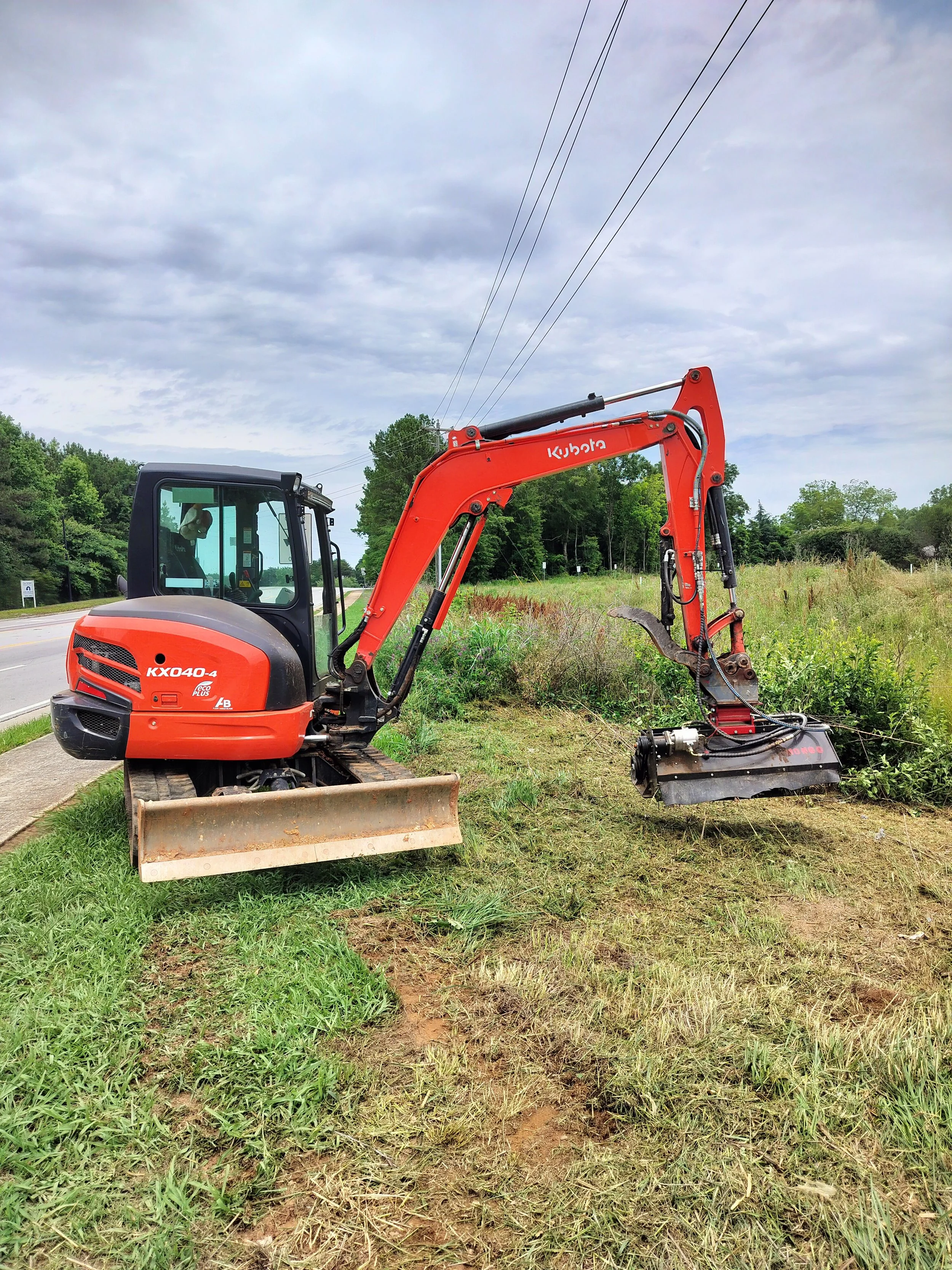 A red Kubota mini excavator working on a patch of grass next to a road with trees in the background and a cloudy sky overhead.
