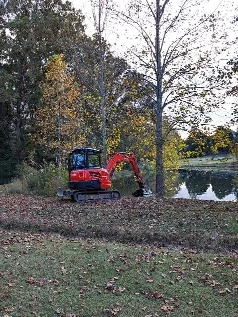 A small red and black excavator is parked on a grassy area near a lake, surrounded by trees with autumn foliage.