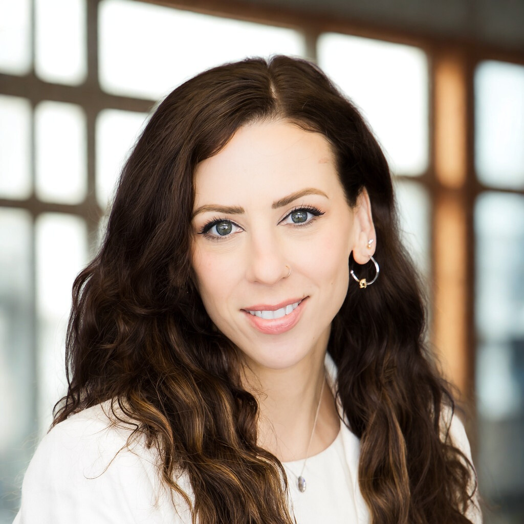 A young woman with long, dark wavy hair, wearing earrings and a necklace, smiles at the camera indoors with large windows in the background.