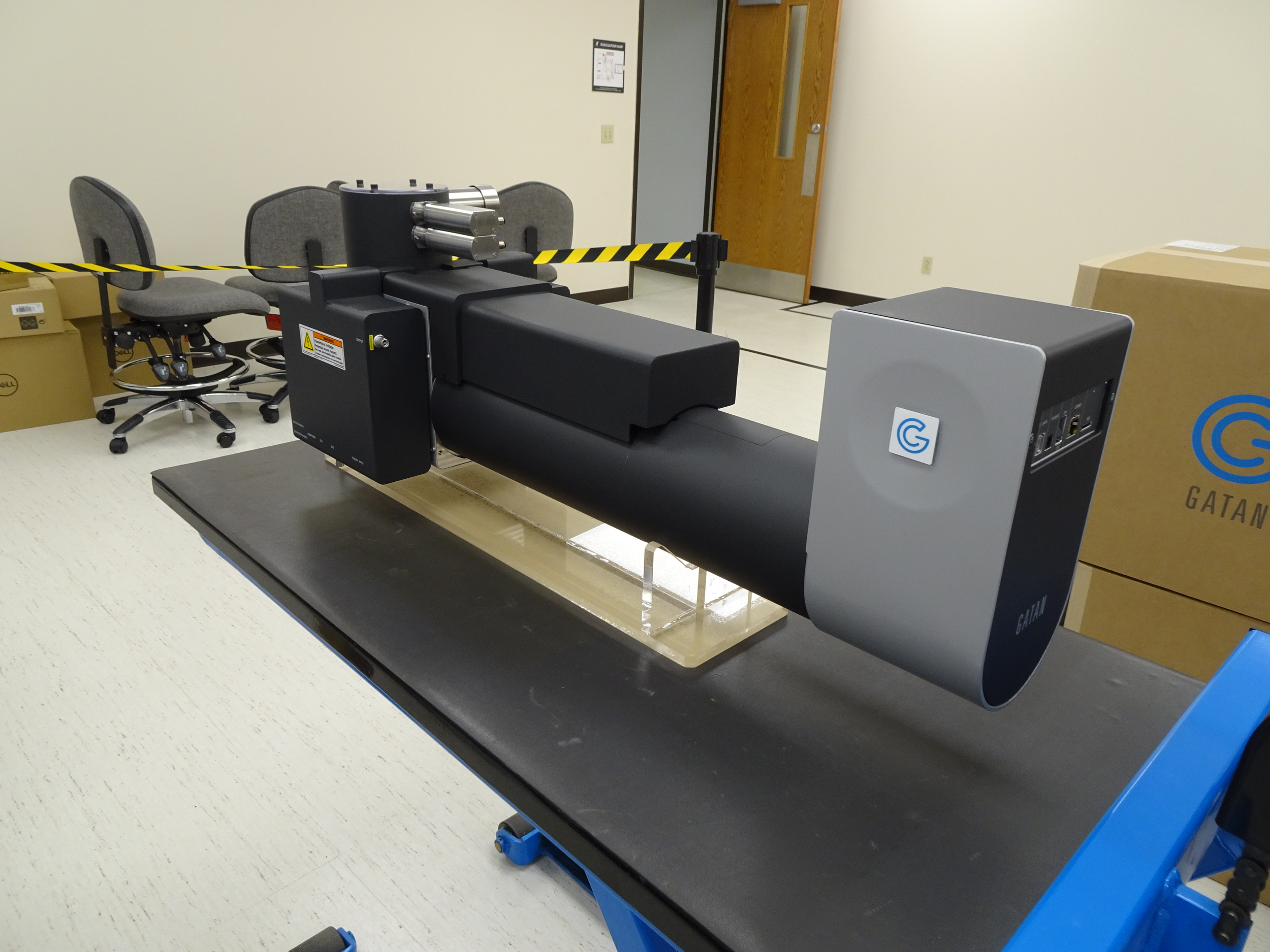 A Gatan cryo-electron microscope in a laboratory setting, with black and gray exterior and a control panel on the side, placed on a black table with a blue support structure.