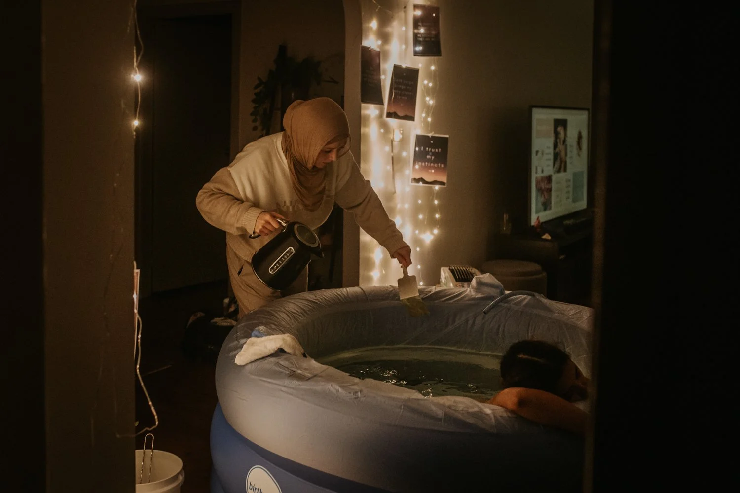 A midwife is pouring water into an inflatable hot tub with a women inside, in a dimly lit room decorated with string lights and inspirational posters on the wall.