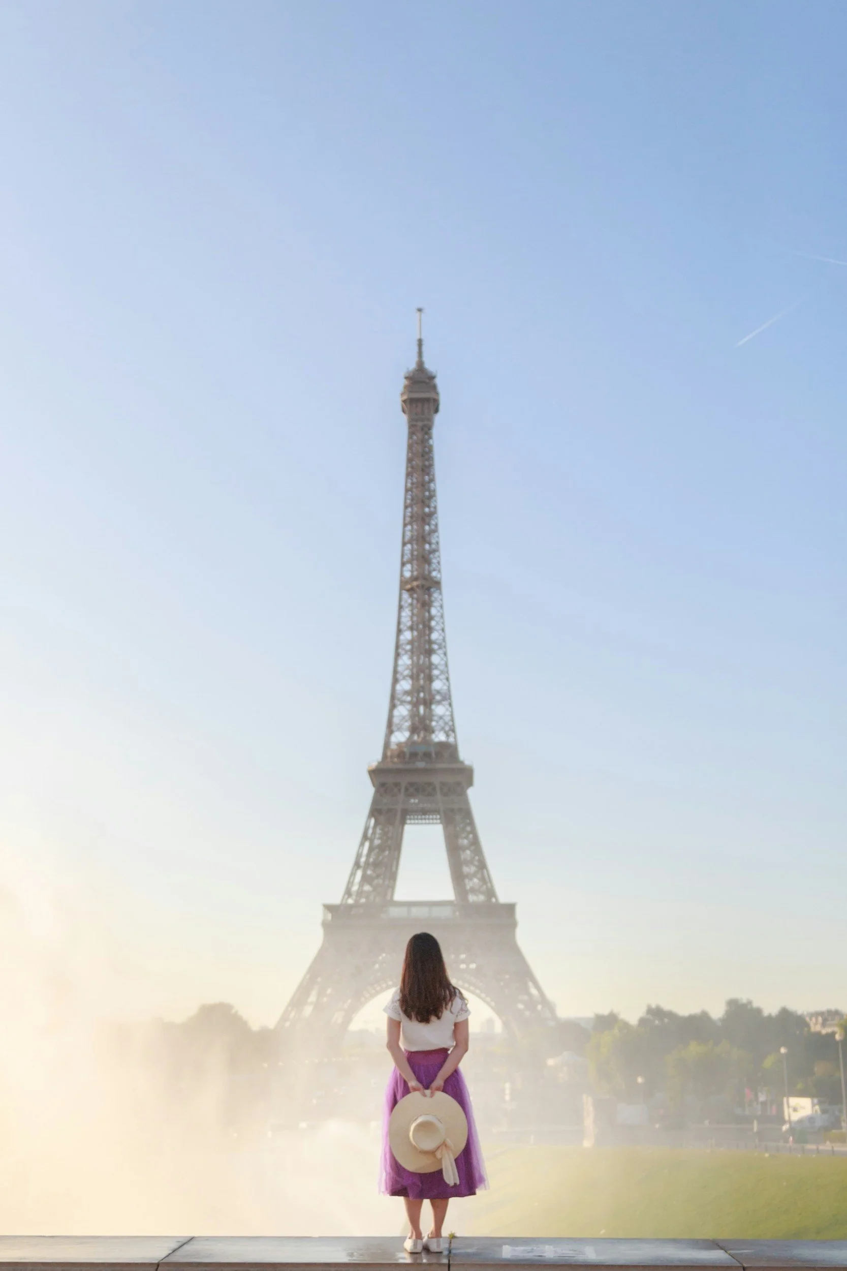 A woman standing with her back to the camera, holding a wide-brimmed hat, looking at the Eiffel Tower in Paris, France.