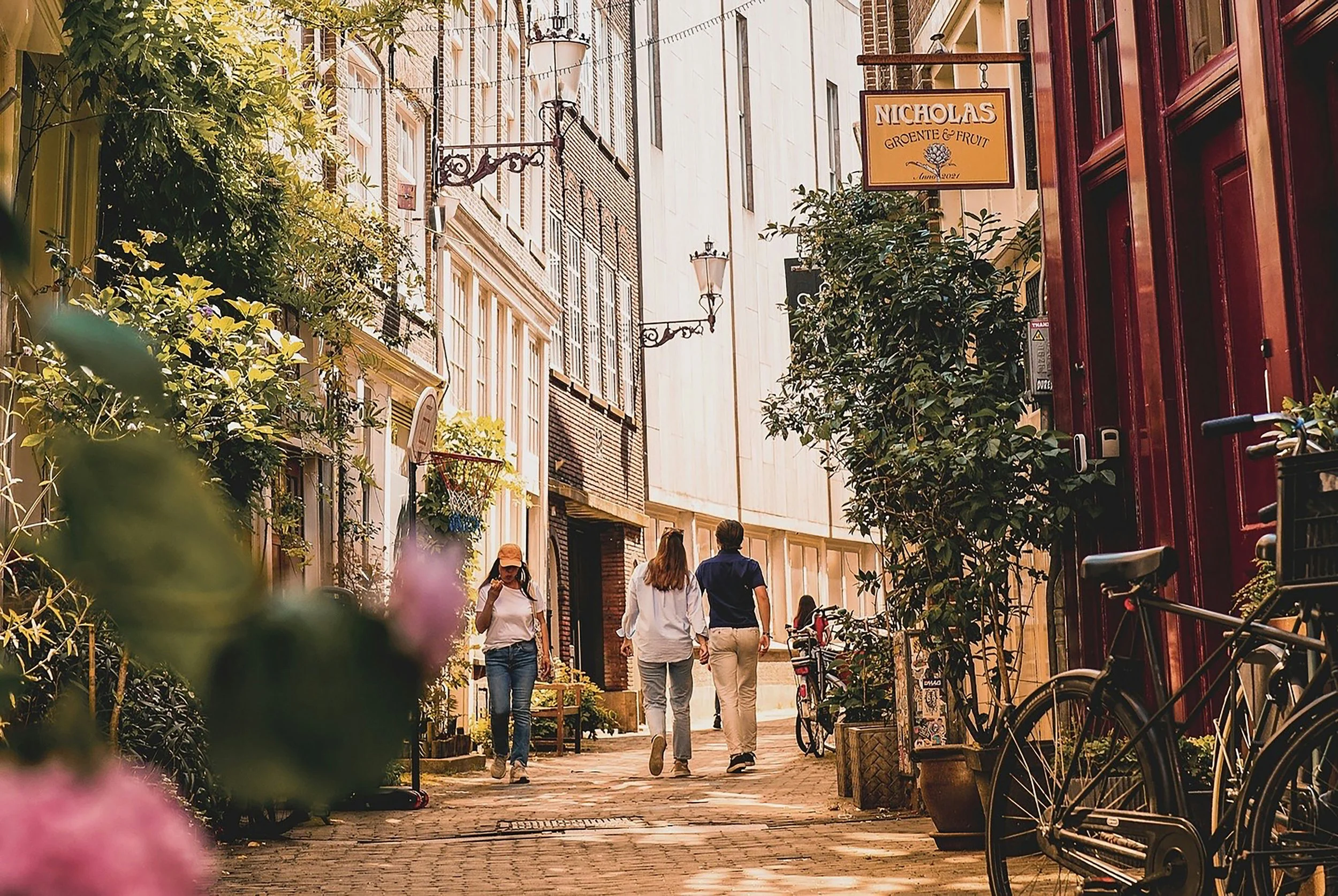 A narrow European street with people walking, surrounded by plants, bicycles, and buildings with warm lighting.