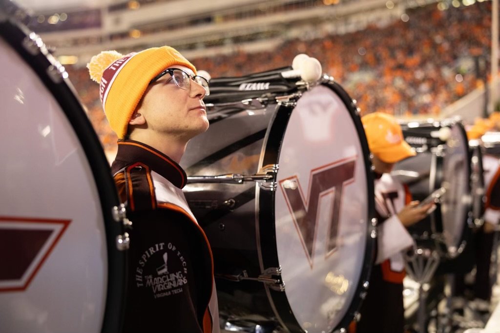 A drummer wearing glasses and a yellow beanie with a pompom, standing behind a bass drum with a Virginia Cavaliers logo, in a stadium during a game.