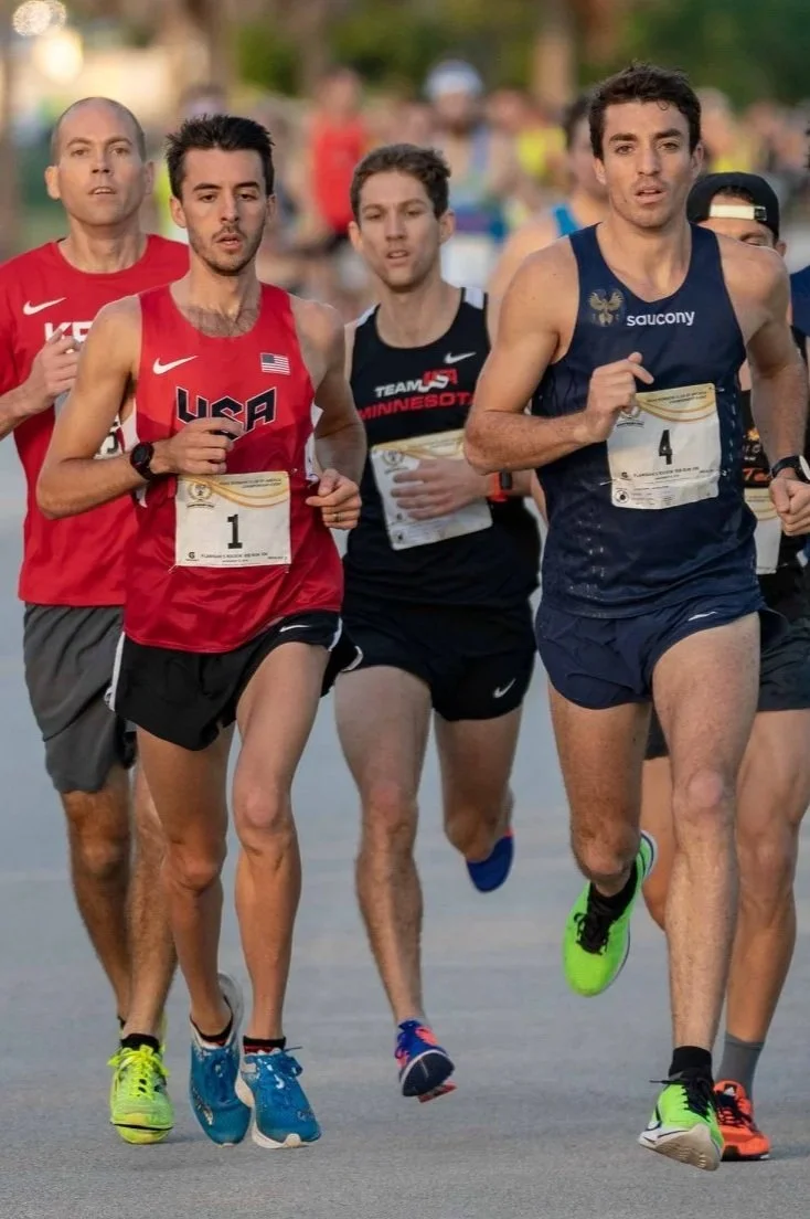 Group of male runners participating in a road race, wearing athletic gear and bib numbers, with trees and other runners in the background.