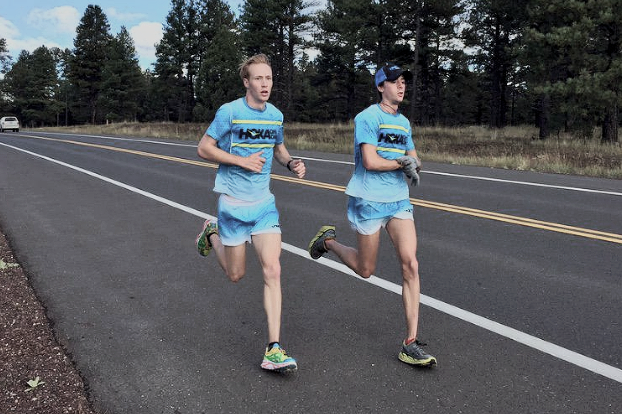 Two young men running on the side of a paved road in a rural area with trees in the background, wearing matching blue running shirts and shorts.