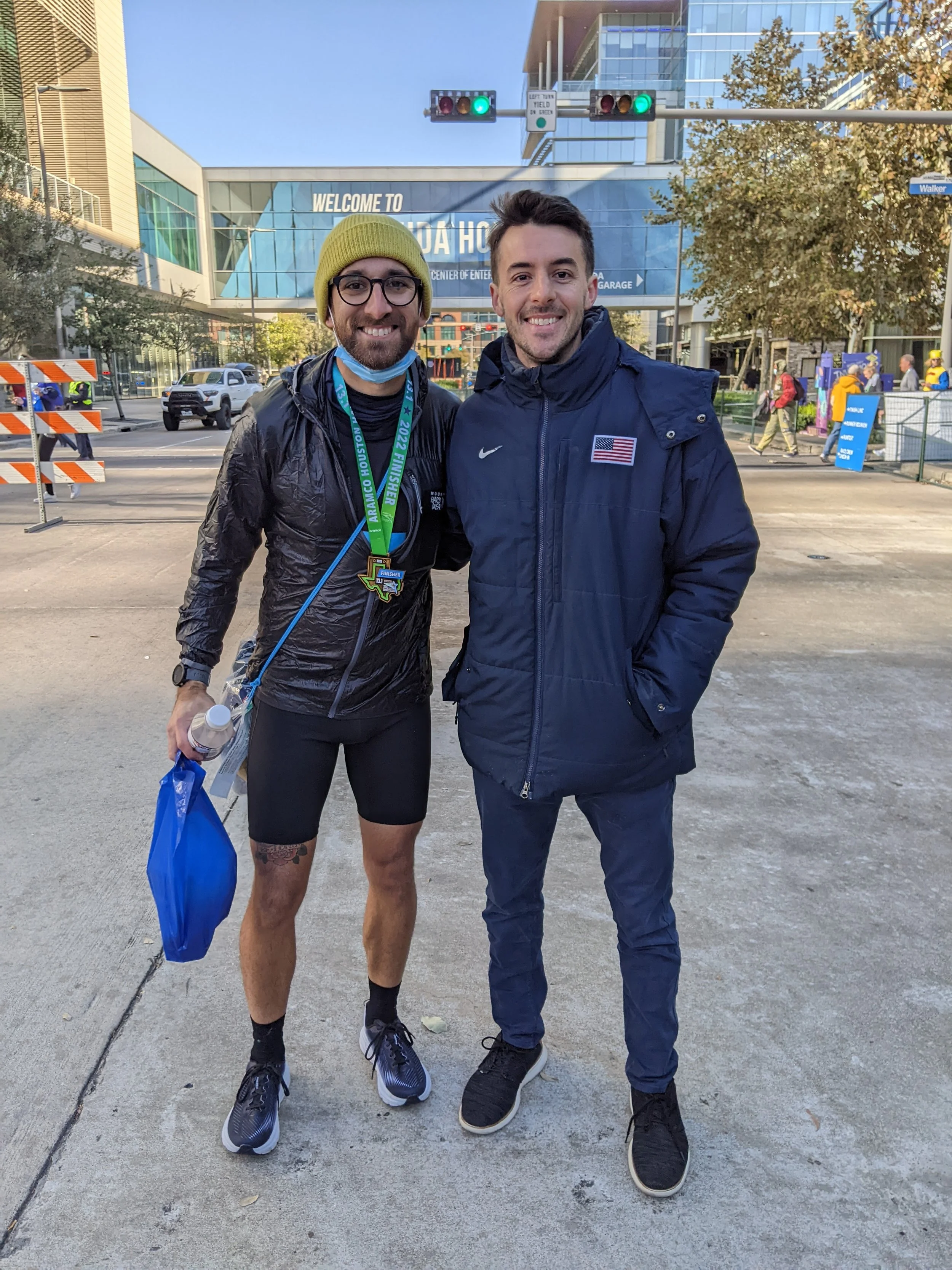 Two men smiling and standing outdoors in front of a building with a sign that says 'Welcome to the NRG Stadium'. One man is dressed in athletic gear with a medal and a blue bag, suggesting he has just finished a race. The other man is wearing a navy jacket with an American flag patch. They are posing together, with traffic lights and trees visible in the background.