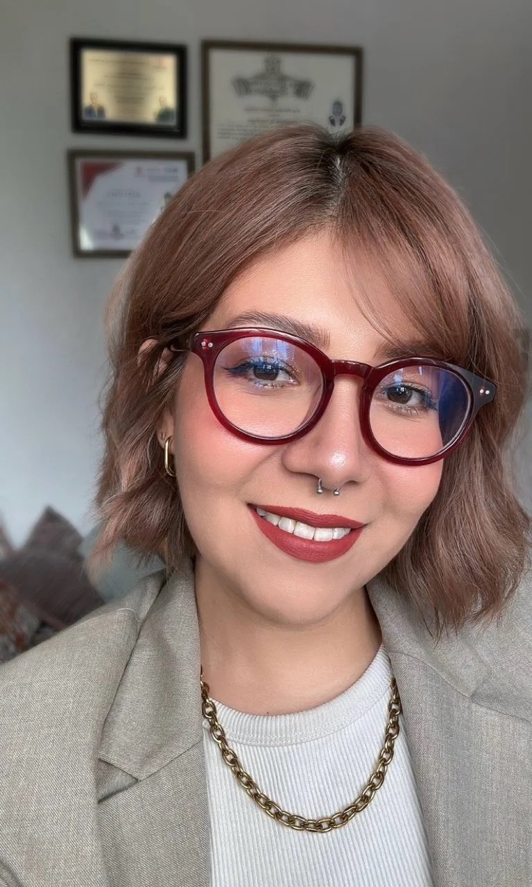 Retrato de una mujer con gafas grandes de color rojo, corte de cabello rubio con ondas, y sonriendo. Ella lleva un abrigo beige, una camiseta blanca, y un collar de cadena gruesa dorada. En el fondo, hay diplomas o certificados colgados en la pared.