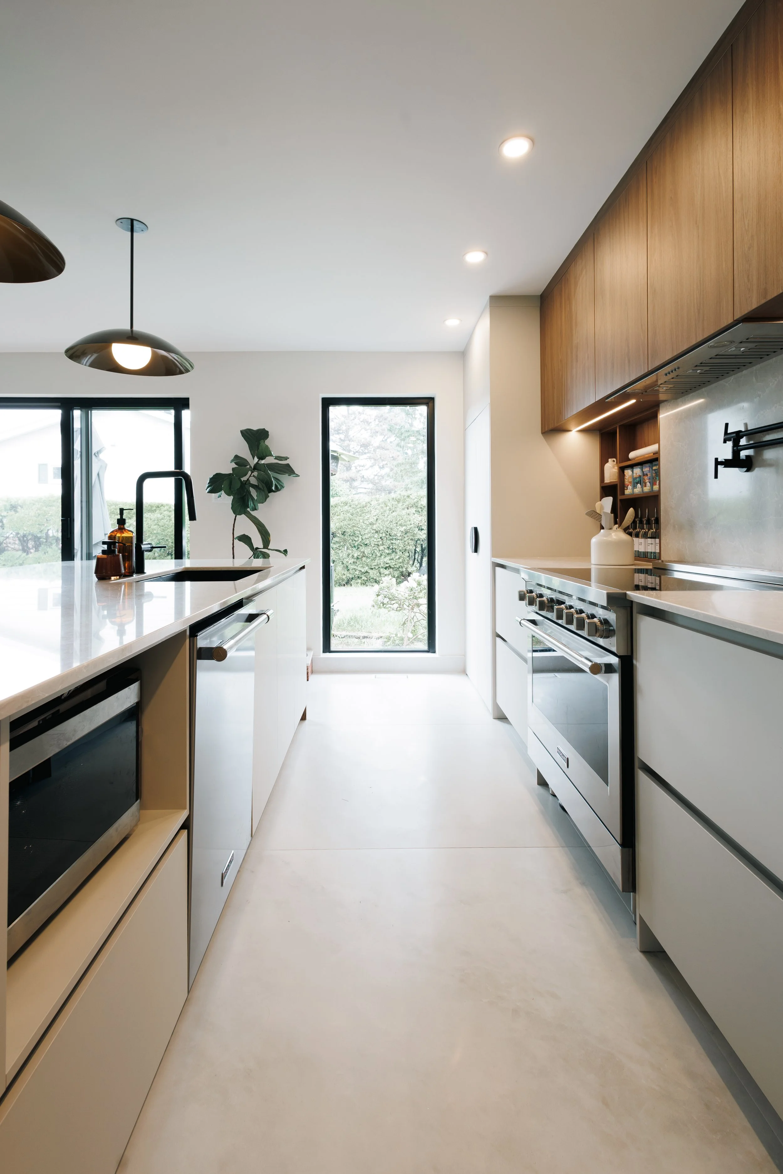Cuisine moderne avec îlot central, meubles en bois clair, appareils en acier inoxydable, grande fenêtre donnant sur un jardin, plante verte sur l'îlot, lampes suspendues noires, murs blancs, sol en béton ou carrelage clair.