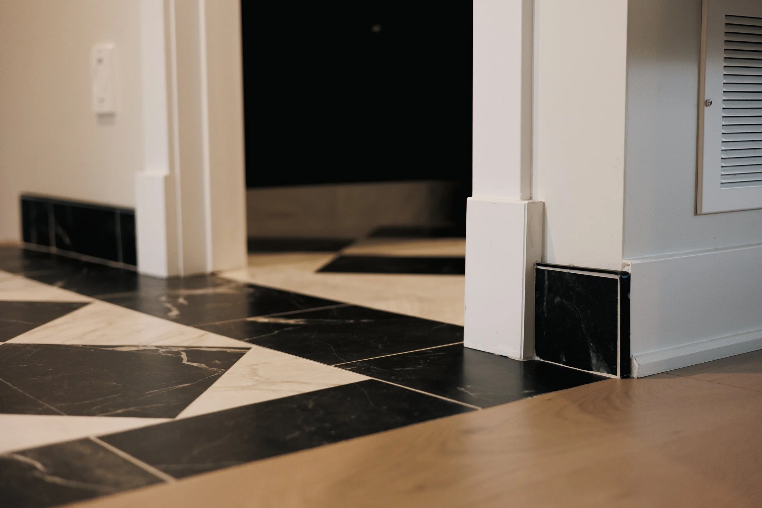 Vue d'un coin de pièce avec un sol en carreaux noirs et blancs, un mur blanc et des appareils électriques encastrés, dont un radiateur électrique gris à l'avant-droite