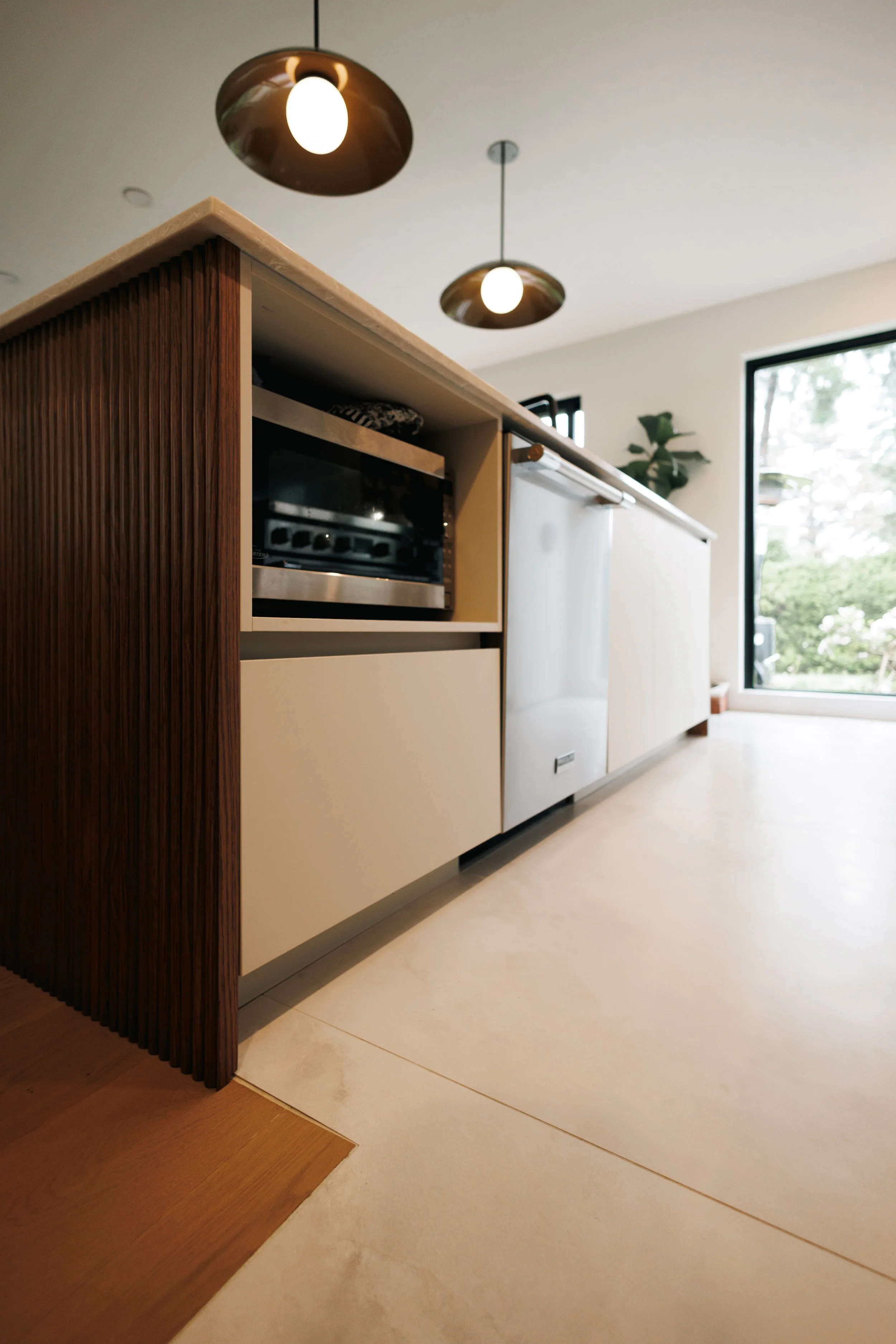 Cliché d'une cuisine moderne avec un plan de travail en bois clair, armoires blanches et un four intégré, avec une grande fenêtre laissant entrer la lumière naturelle, un luminaire suspendu au plafond.
