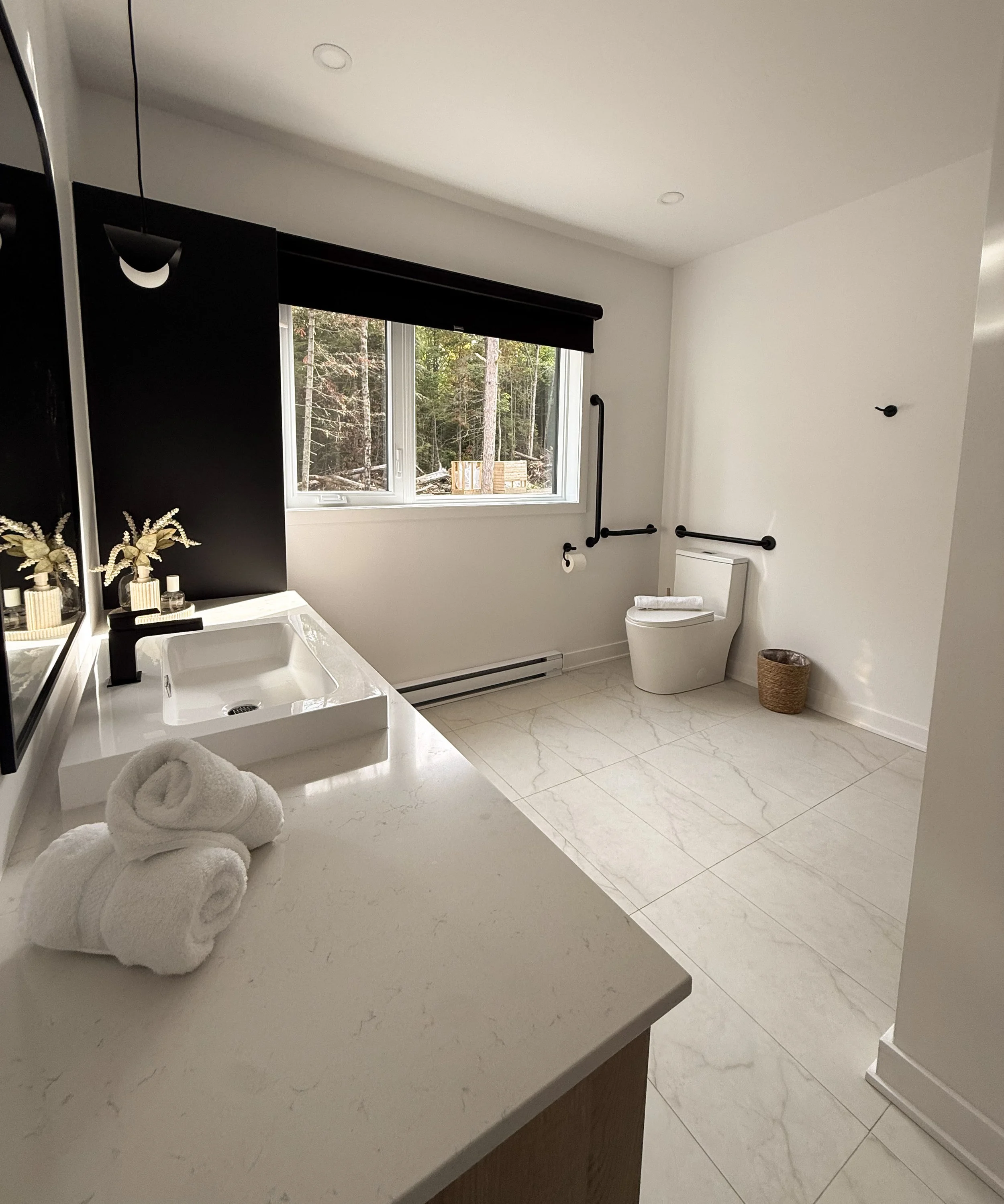 Salle de bain moderne avec un lavabo carré blanc, des serviettes roulées en haut de la commode, une fenêtre donnant sur une forêt, un toilet à côté, et des accessoires en noir et en jute.
