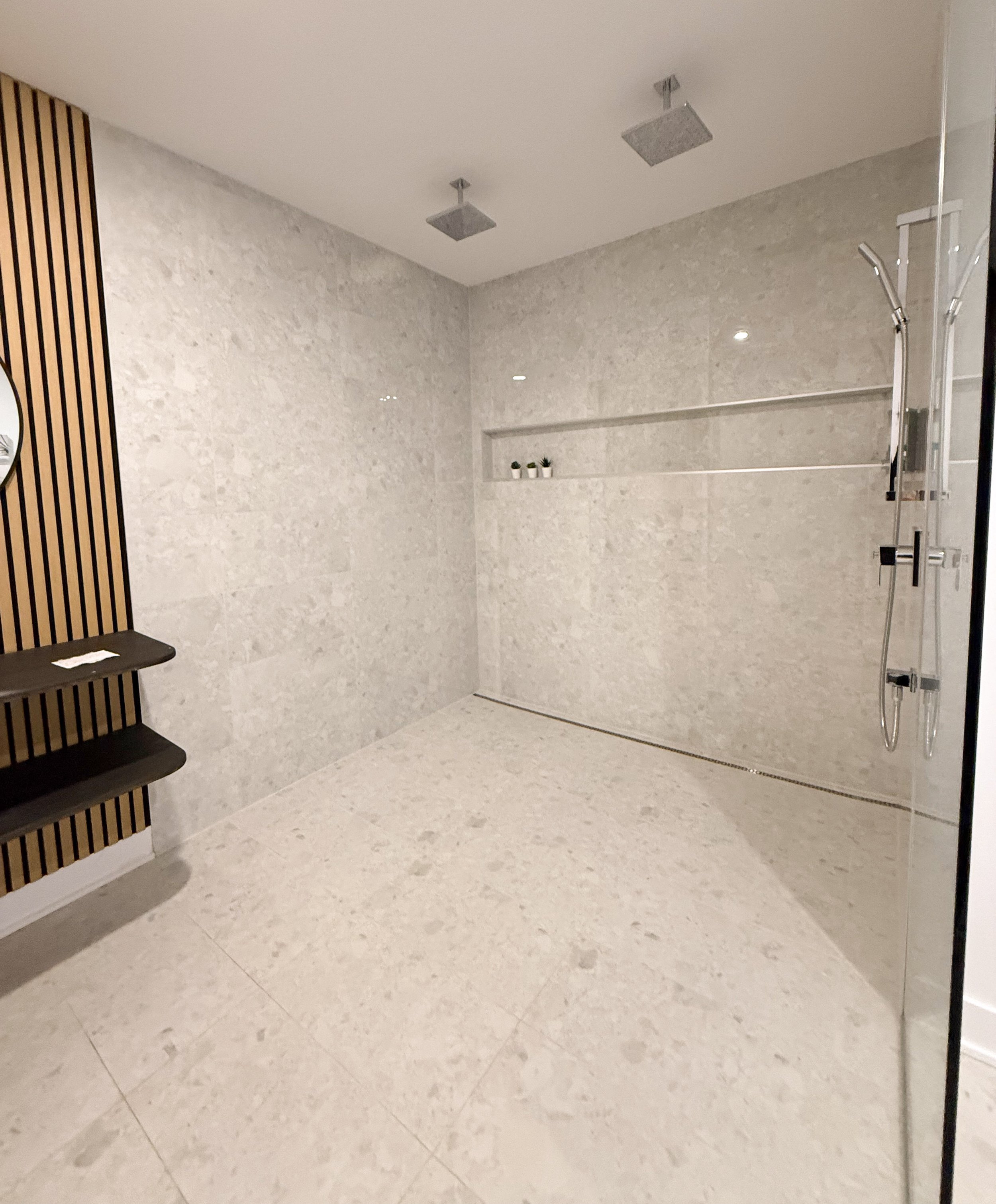 Salle de douche moderne avec murs en pierre claire, deux plafonniers, une étagère intégrée décorée de petites plantes, et un système de douche à main avec deux têtes de douche.