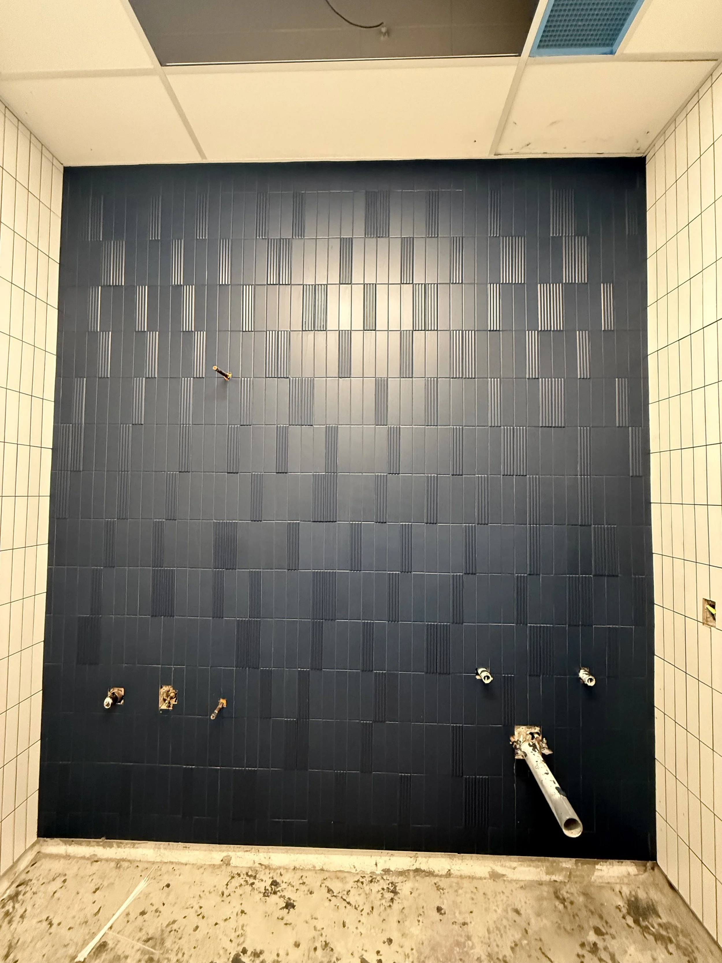 Un mur de salle de bain en cours de rénovation avec des carreaux noirs et blancs, plomberie à l'avant, et du sol sale avec des débris.