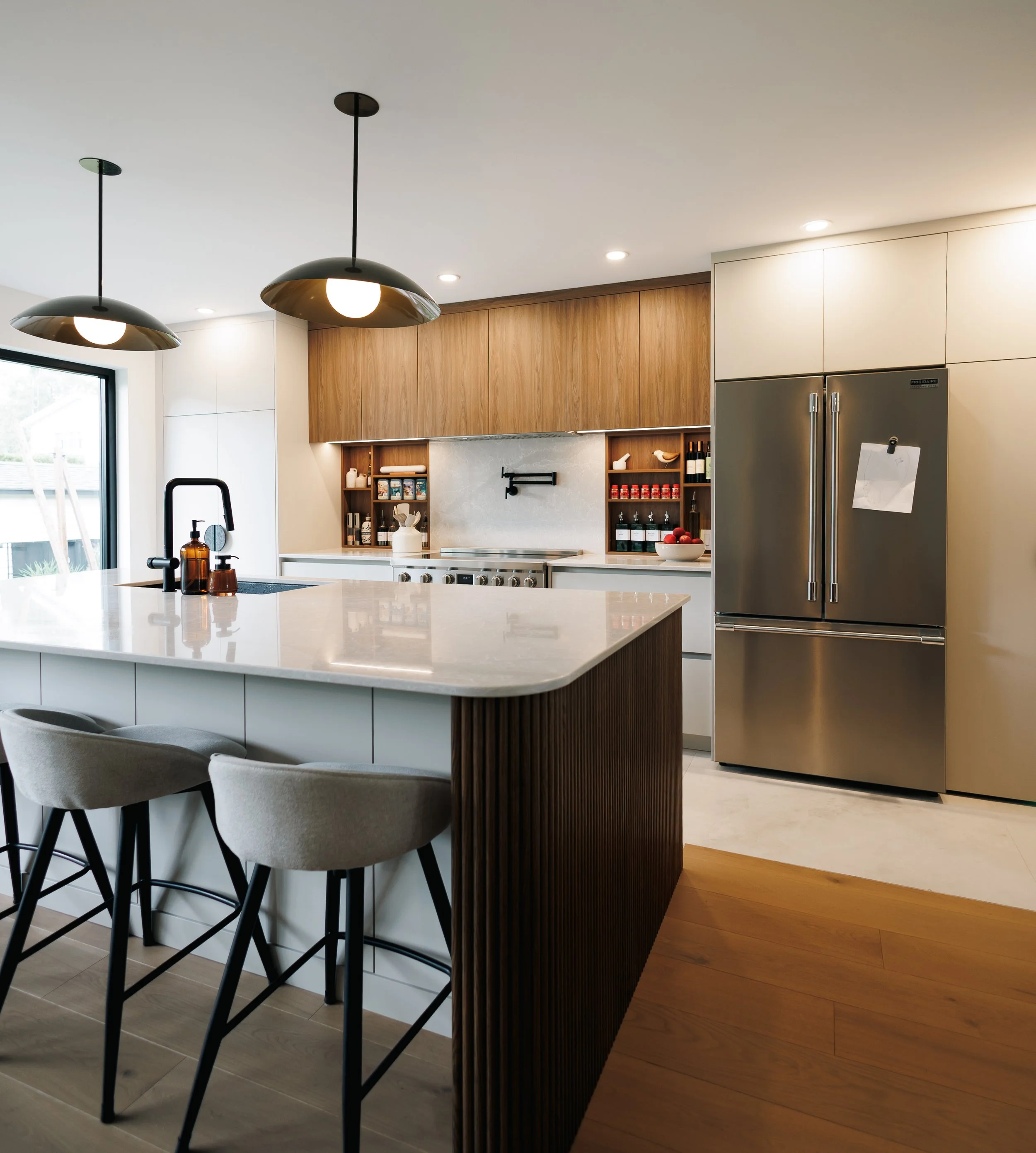 Cuisine moderne avec un îlot central en pierre blanche, luminaires suspendus noirs, armoires en bois clair, appareils en acier inoxydable, et un coin repas avec des chaises élégantes.