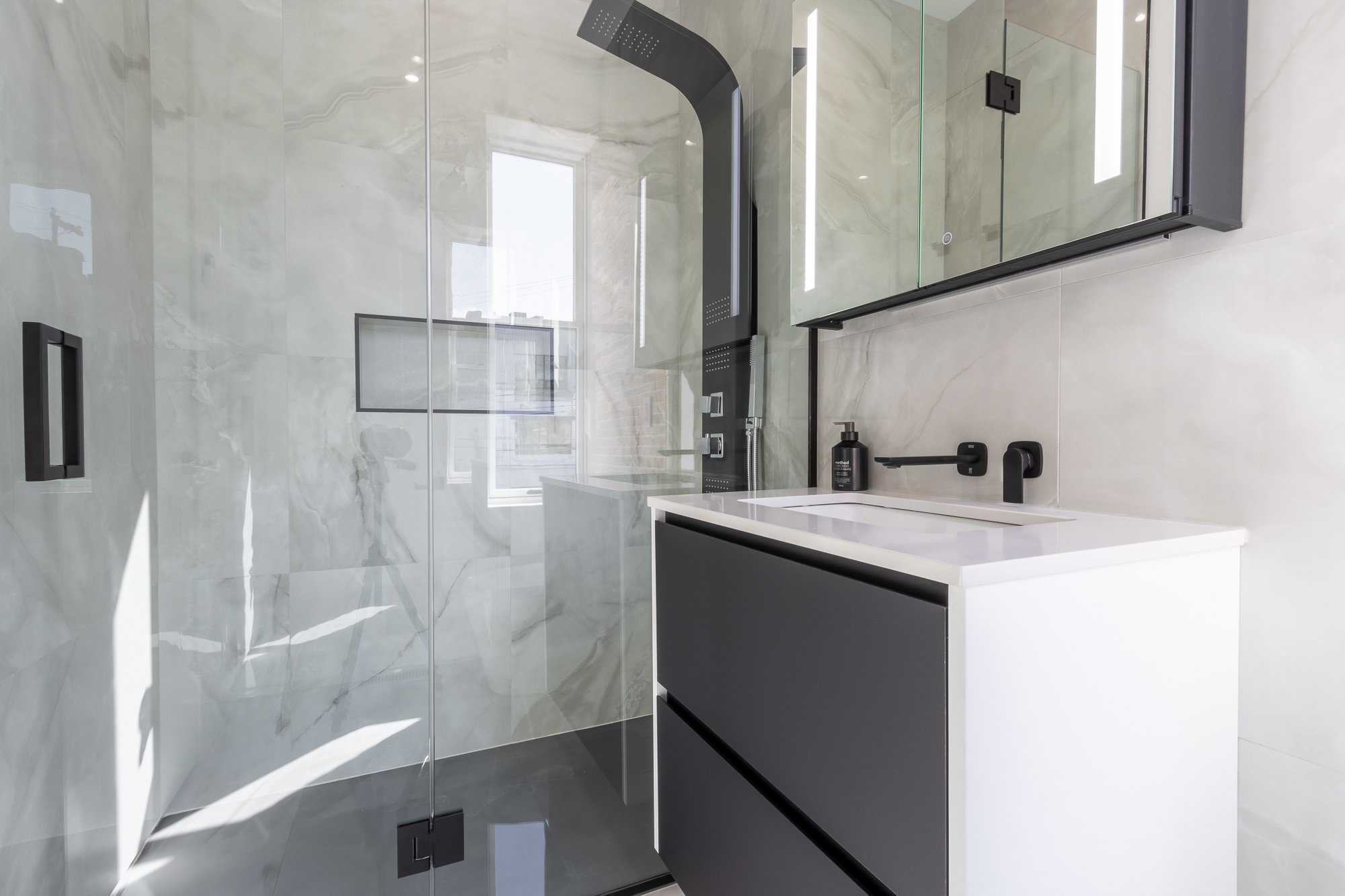 Salle de bain moderne avec douche à l'italienne en verre, meuble de lavabo noir et blanc, miroir mural avec éclairage intégré, murs en marbre clair et accessoires noirs.