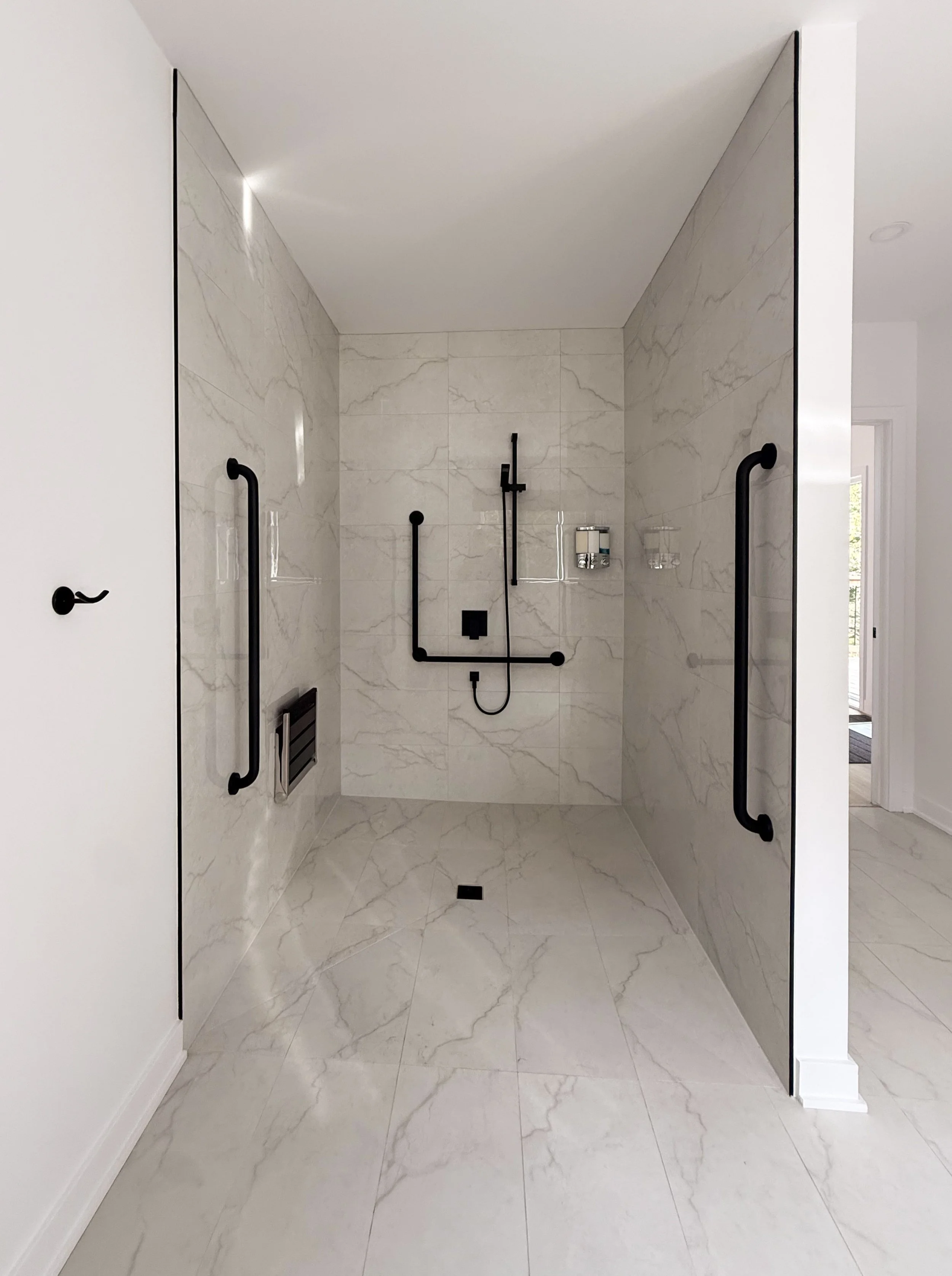 Salle de bain avec murs et sol en marbre clair, deux portes en verre avec poignées noires, une douche à l'italienne équipée d'une barre de maintien noire, un porte-savon et une grilles de ventilation, une porte d'entrée ouverte avec vue sur une pièce