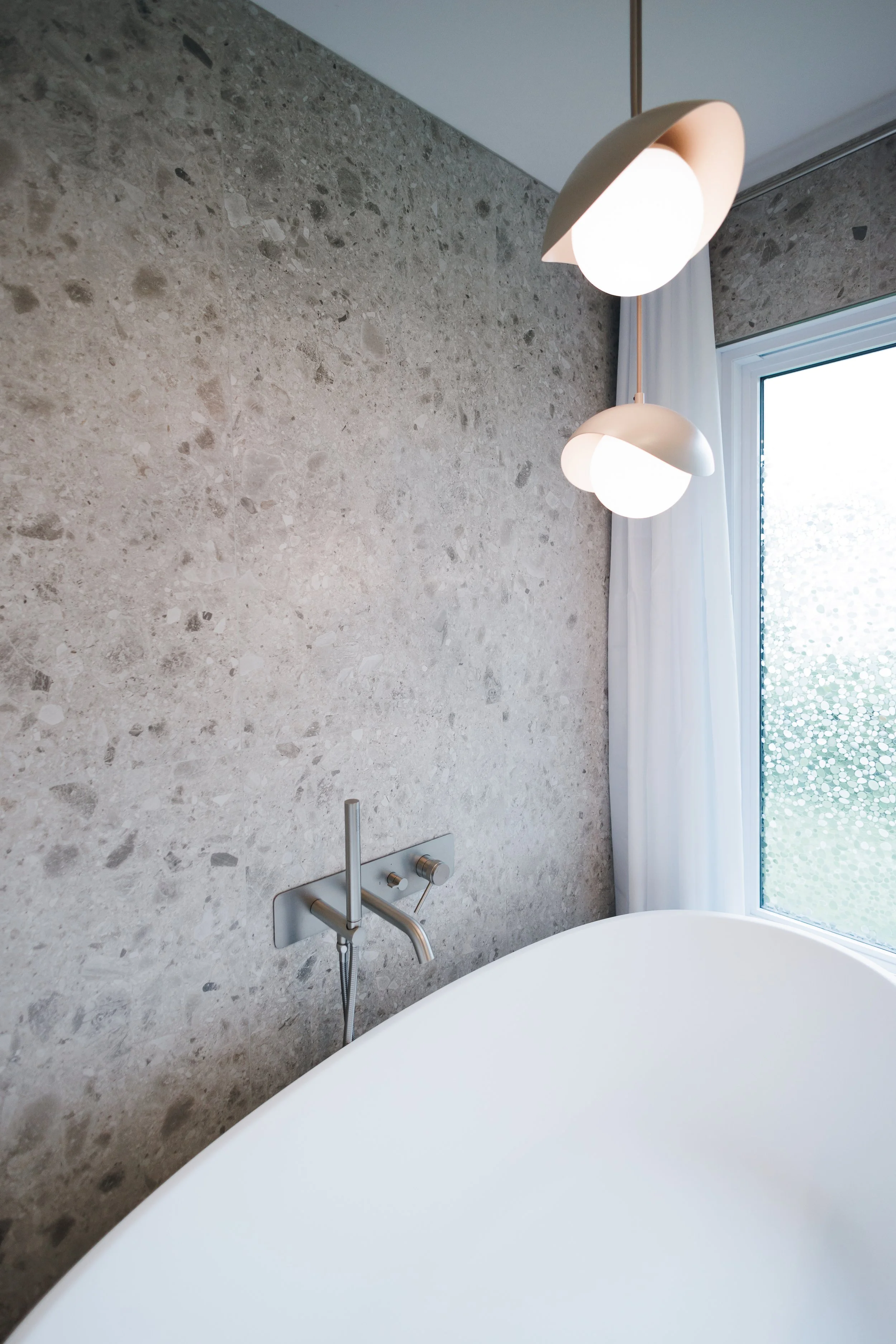 Une baignoire blanche avec un mur en pierre grise derrière. Il y a une fenêtre avec un rideau blanc, et un éclairage moderne suspendu au plafond.