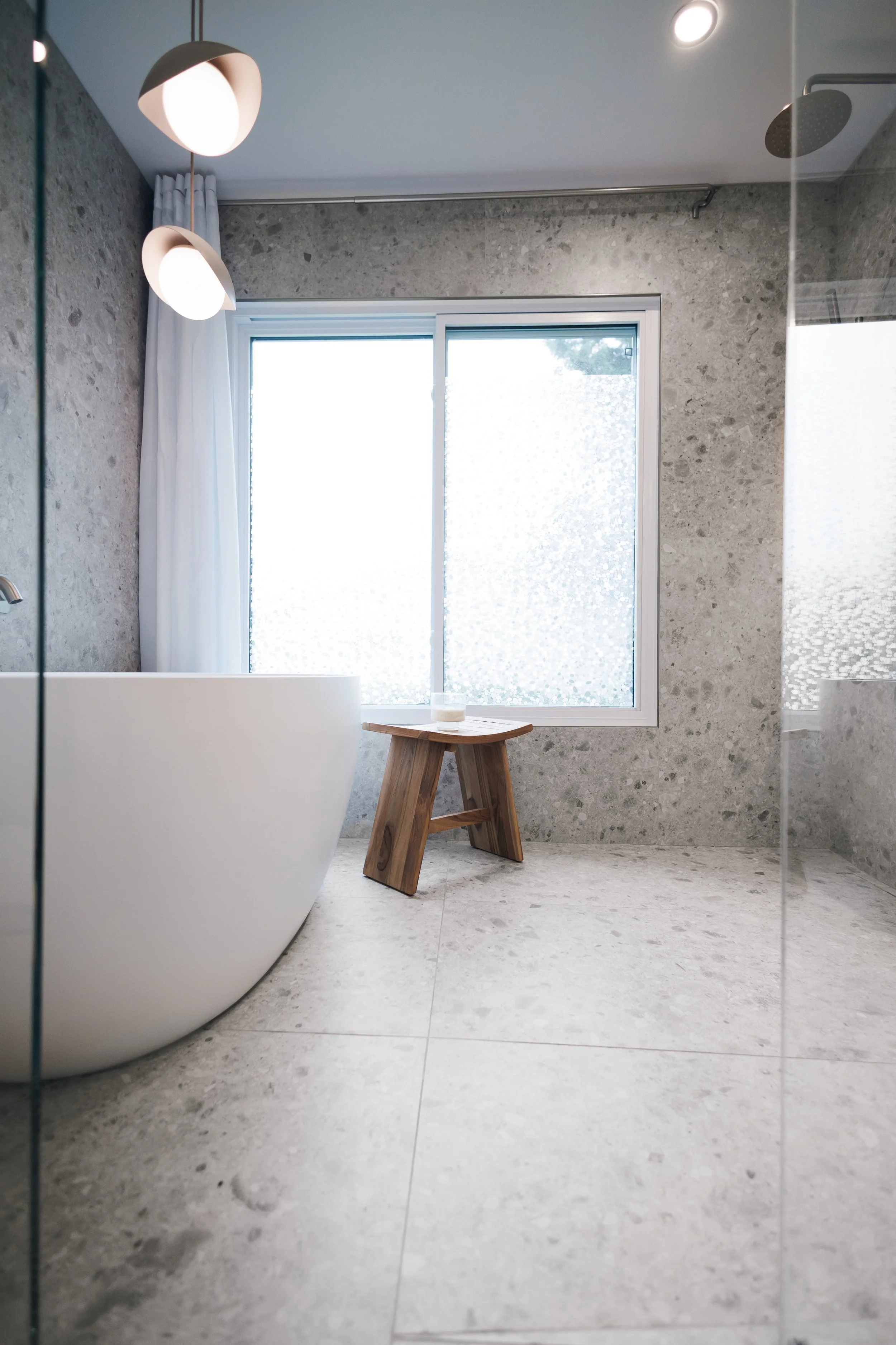 Salle de bain avec une baignoire en forme de coquillage blanc, mur en pierre grise, fenêtre avec rideau blanc, petit banc en bois, lumière encastrée au plafond.