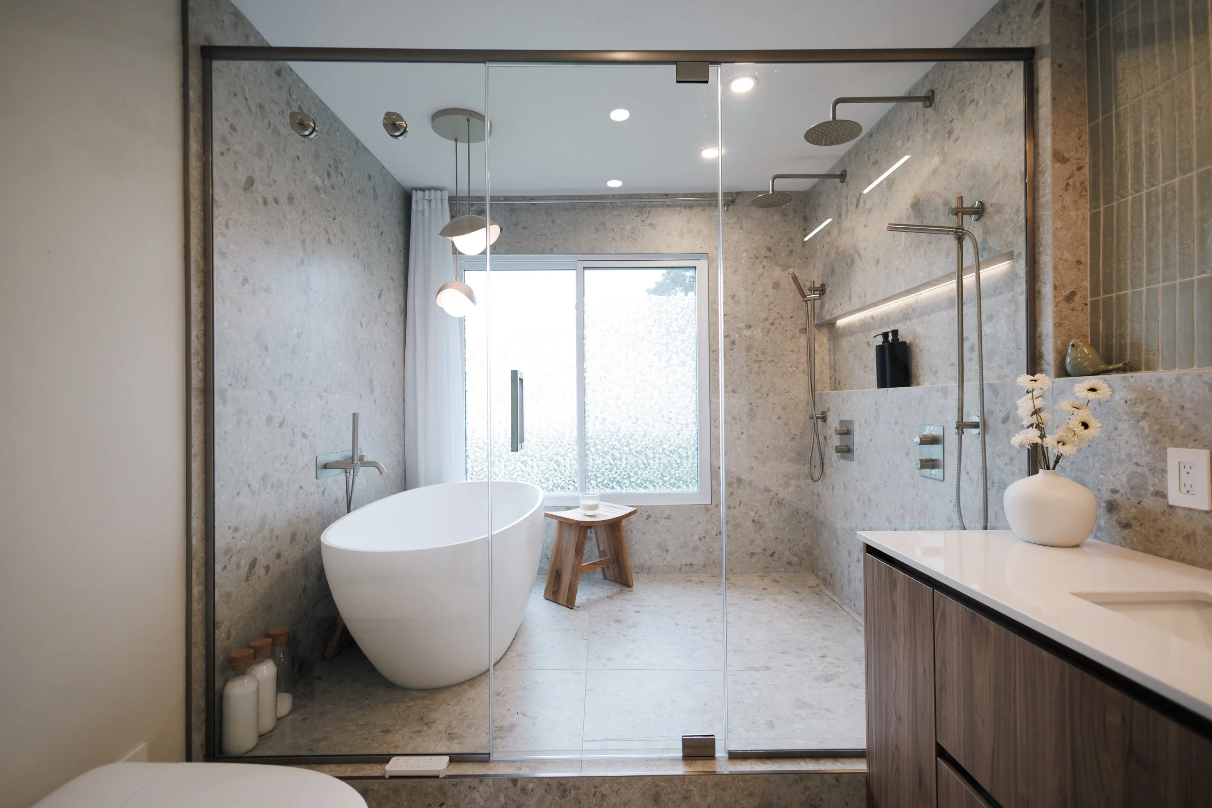 Salle de bain moderne avec baignoire autoportante, douche à l'italienne avec deux pommeaux de douche, fenêtre laissant entrer la lumière naturelle, décoration minimaliste avec des fleurs dans un vase blanc.