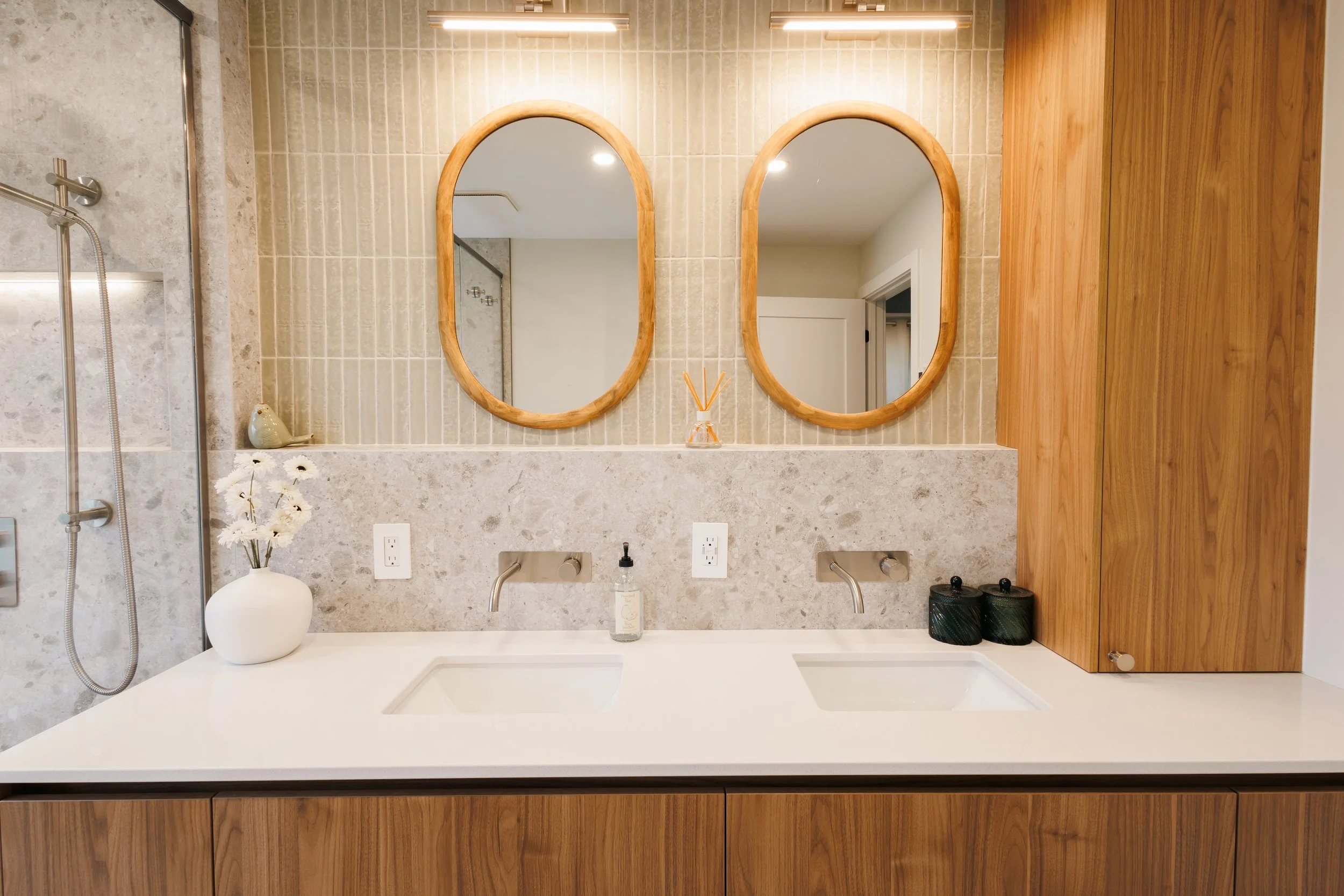 Lavabo double avec deux miroirs ovales en bois au-dessus, décorations florales dans un vase blanc, toilettage moderne, panneau en bois foncé à droite, mur en carrelage beige, éclairage au plafond.
