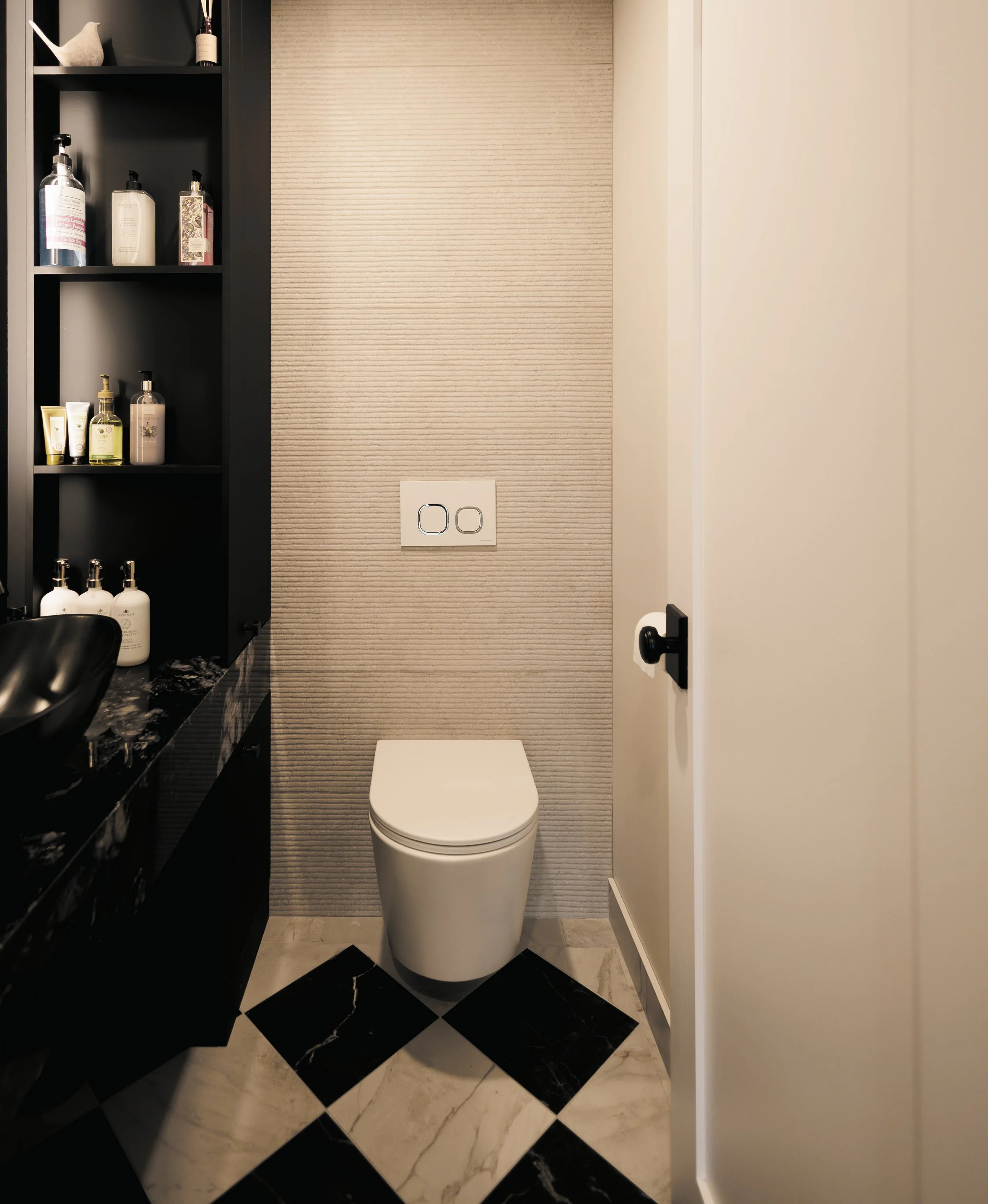 Toilettes avec papier peint beige derrière, avec une douche noire sur la gauche contenant des bouteilles de produits de toilette, et une porte blanche sur la droite.