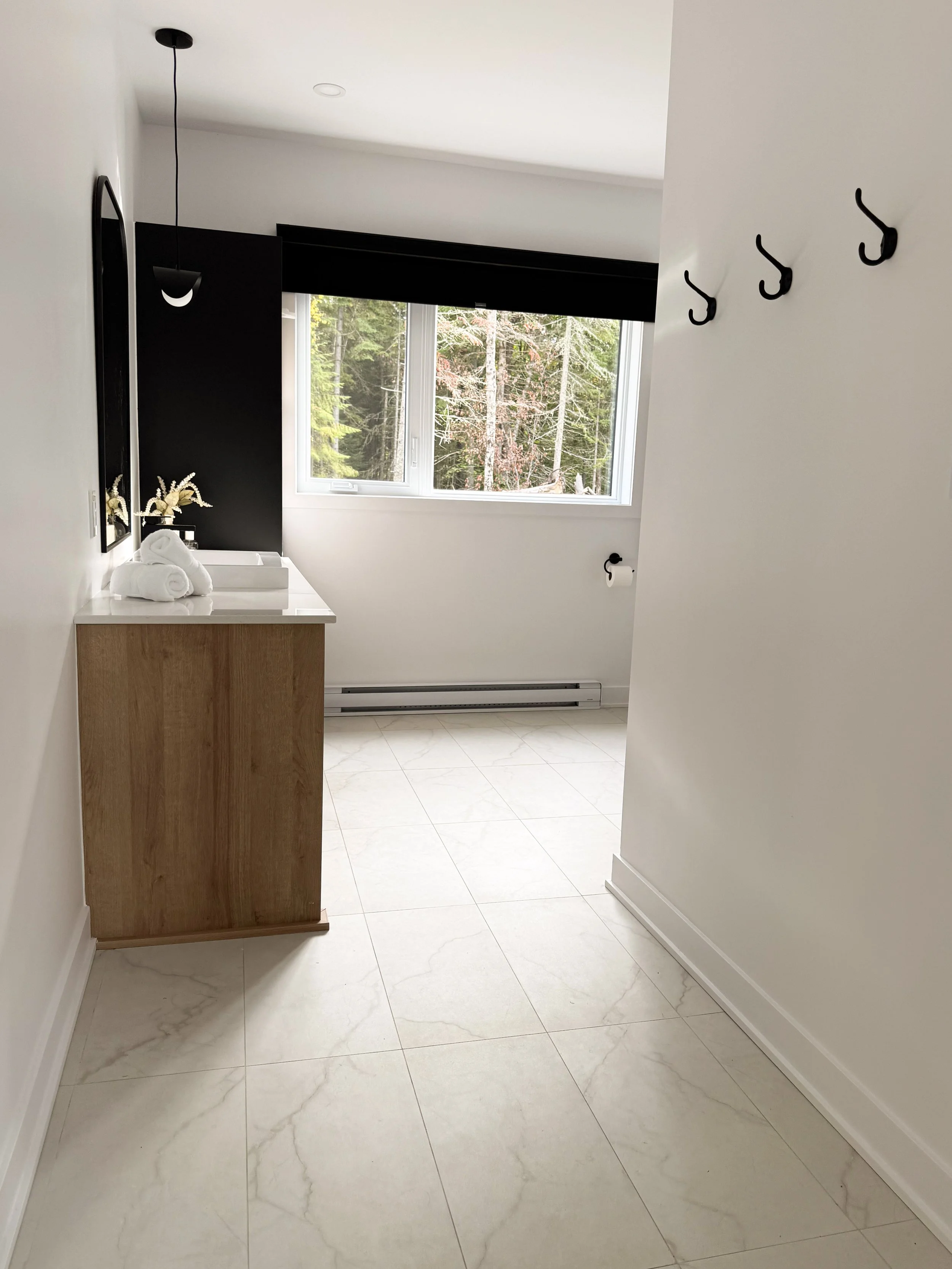 Une pièce de salle de bain moderne avec un meuble en bois, un lavabo blanc, des serviettes roulées, une décoration florale, une fenêtre laissant entrer la lumière naturelle, un mur noir derrière la fenêtre, et un sol en carrelage blanc avec des motif