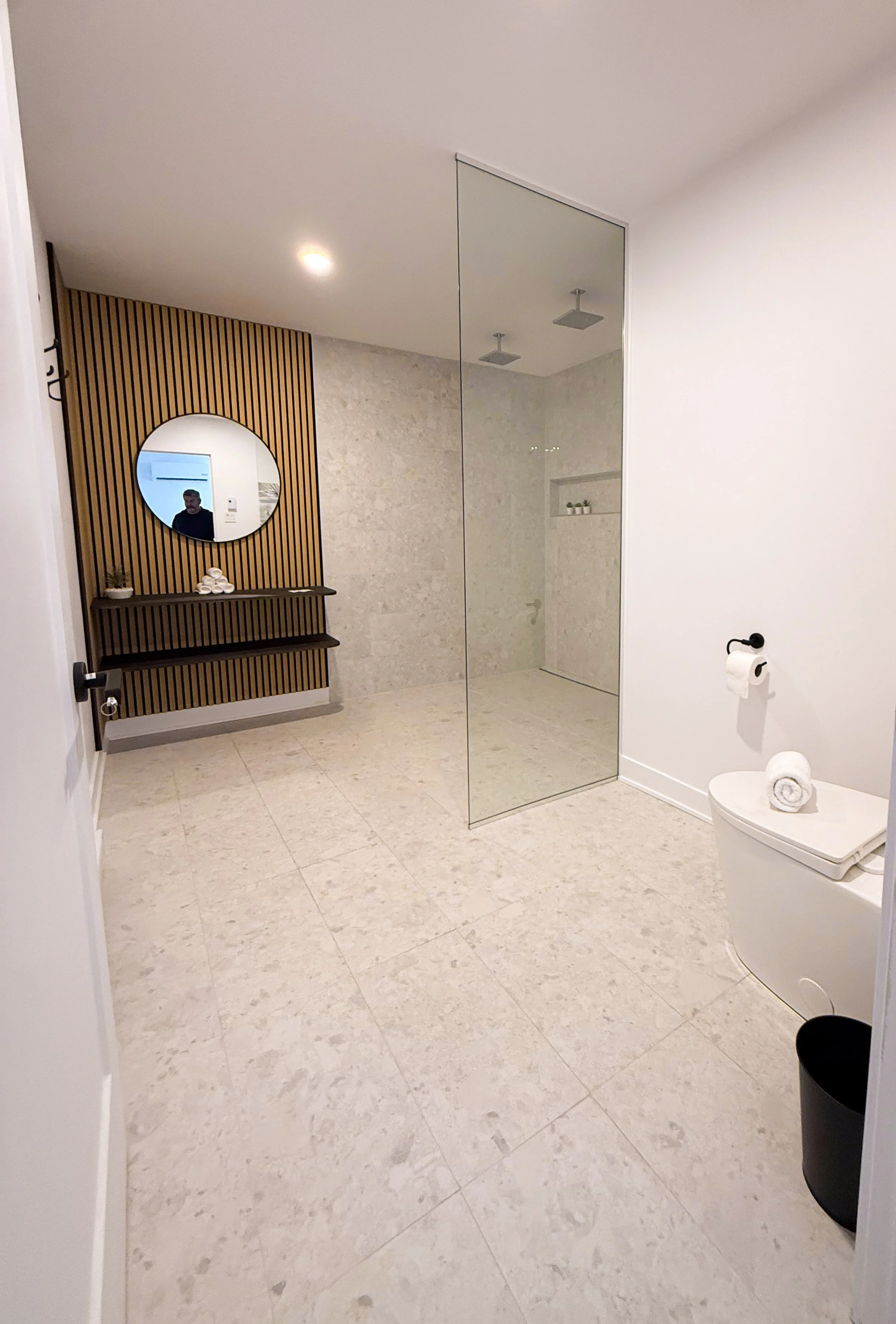 Salle de bain moderne avec sol en pierre, mur en pierre beige, miroir rond au mur en bois rayé, zone de douche séparée avec parois en verre, et toilette avec rouleau de papier toilette