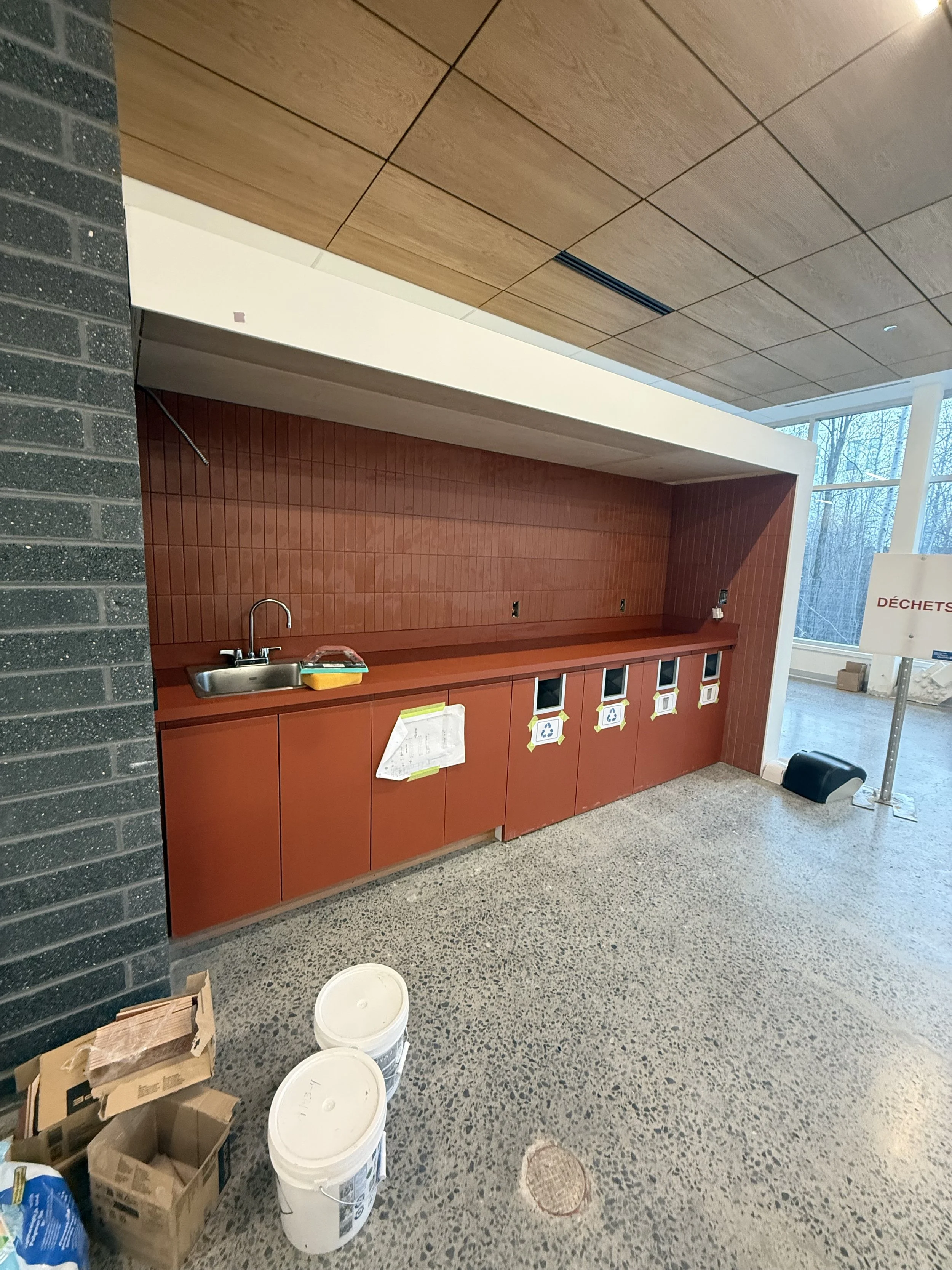 Un espace de cuisine ou de service avec un plan de travail rouge, plusieurs poubelles blanches, des cartons en dessous et à côté, et un grand mur de fenêtres à droite avec des arbres visibles à l'extérieur. Il y a également un panneau indiquant 'DÉCH