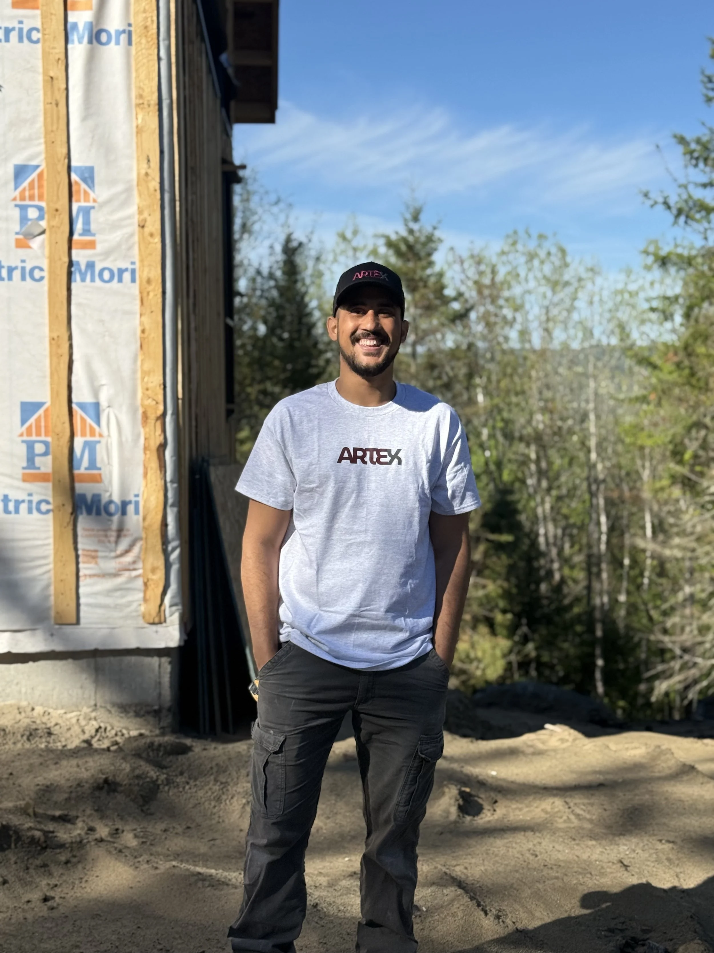 Un homme souriant portant un t-shirt blanc avec le mot 'ARTEX' et une casquette noire, posant à côté d'une maison en construction avec des murs en bois et en matériaux d'isolation, dans un environnement extérieur avec des arbres et un ciel bleu.