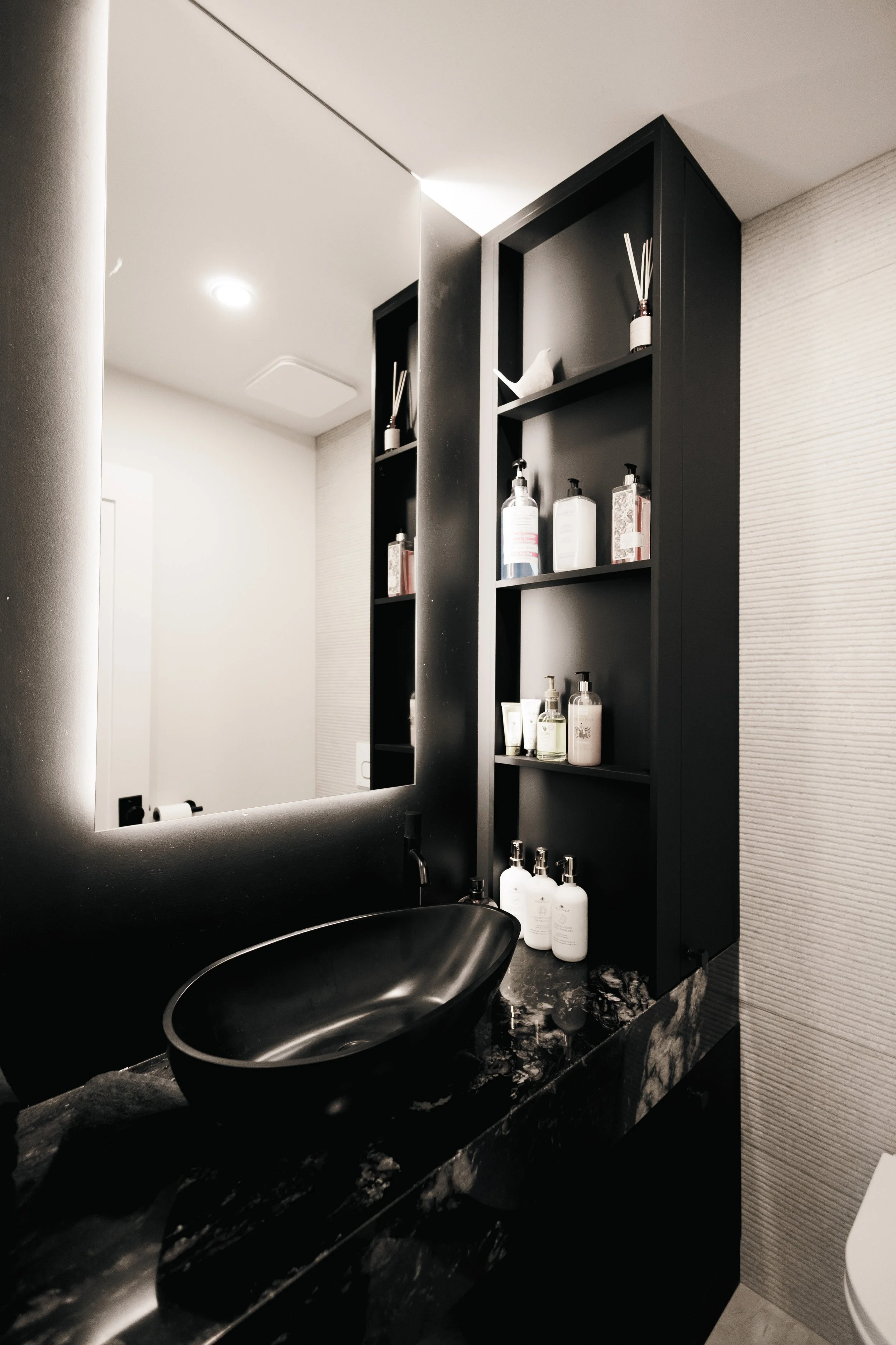 Un miroir au-dessus d'un lavabo noir enuvré d'une planche en pierre avec des bouteilles de produits de toilette, une étagère noire avec des récipients et un diffuseur d'arômes dans une salle de bain moderne.