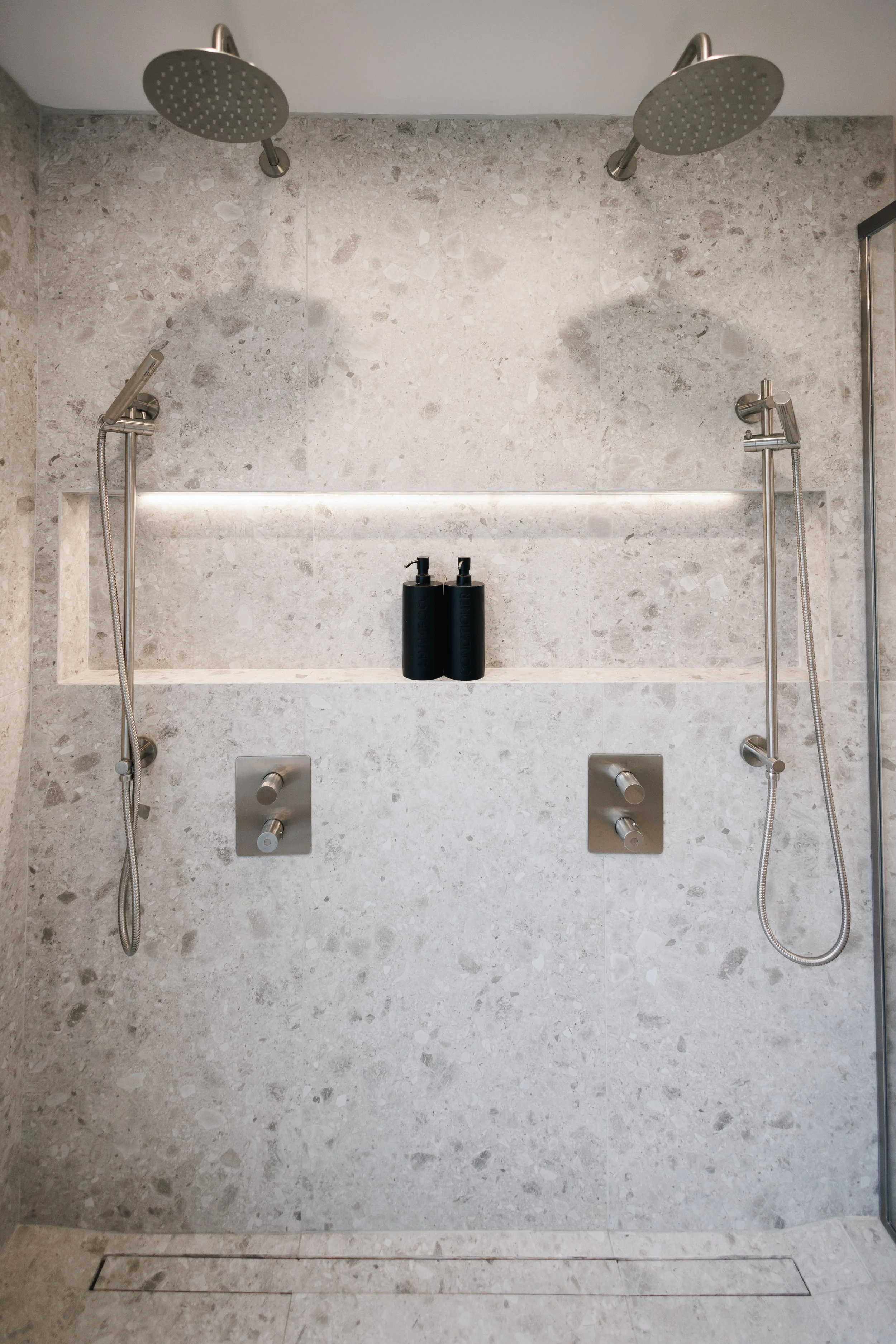 Douche avec deux pommeaux au plafond, deux mitigeurs, deux têtes de douche à main, deux bouteilles de shampooing ou gel douche noires sur une étagère, paroi de douche en verre, sol en pierre avec grille d'évacuation, mur en pierre beige