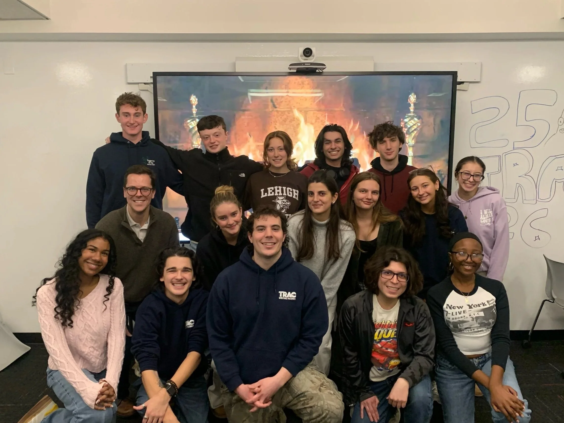 Group of teenage students and adults posing in a classroom in front of a TV screen showing a fireplace scene, with whiteboard and wall art around.