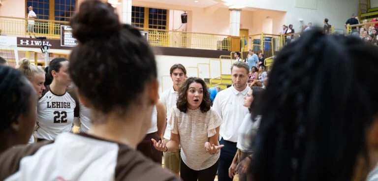 A volleyball coach speaking to her team in a gymnasium during a game or practice, with players listening attentively.