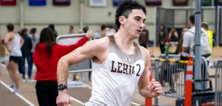 Male athlete running indoors wearing a white tank top with "LEHLL" printed on it, surrounded by people and track and field equipment.