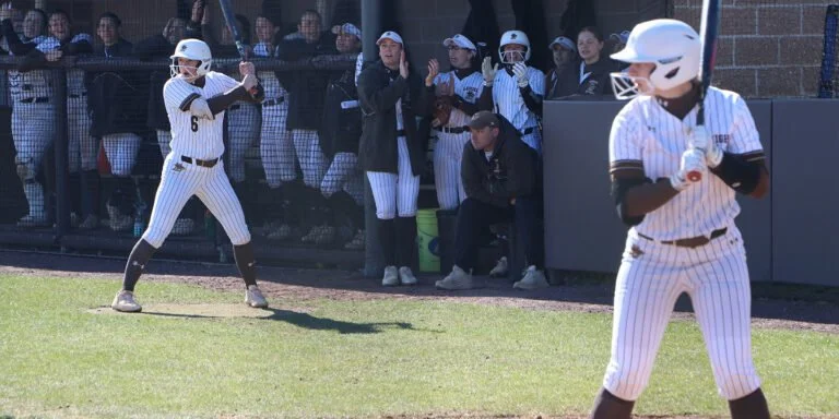 Youth baseball players in striped uniforms and protective helmets on the field, with spectators and coaches watching from the dugout.