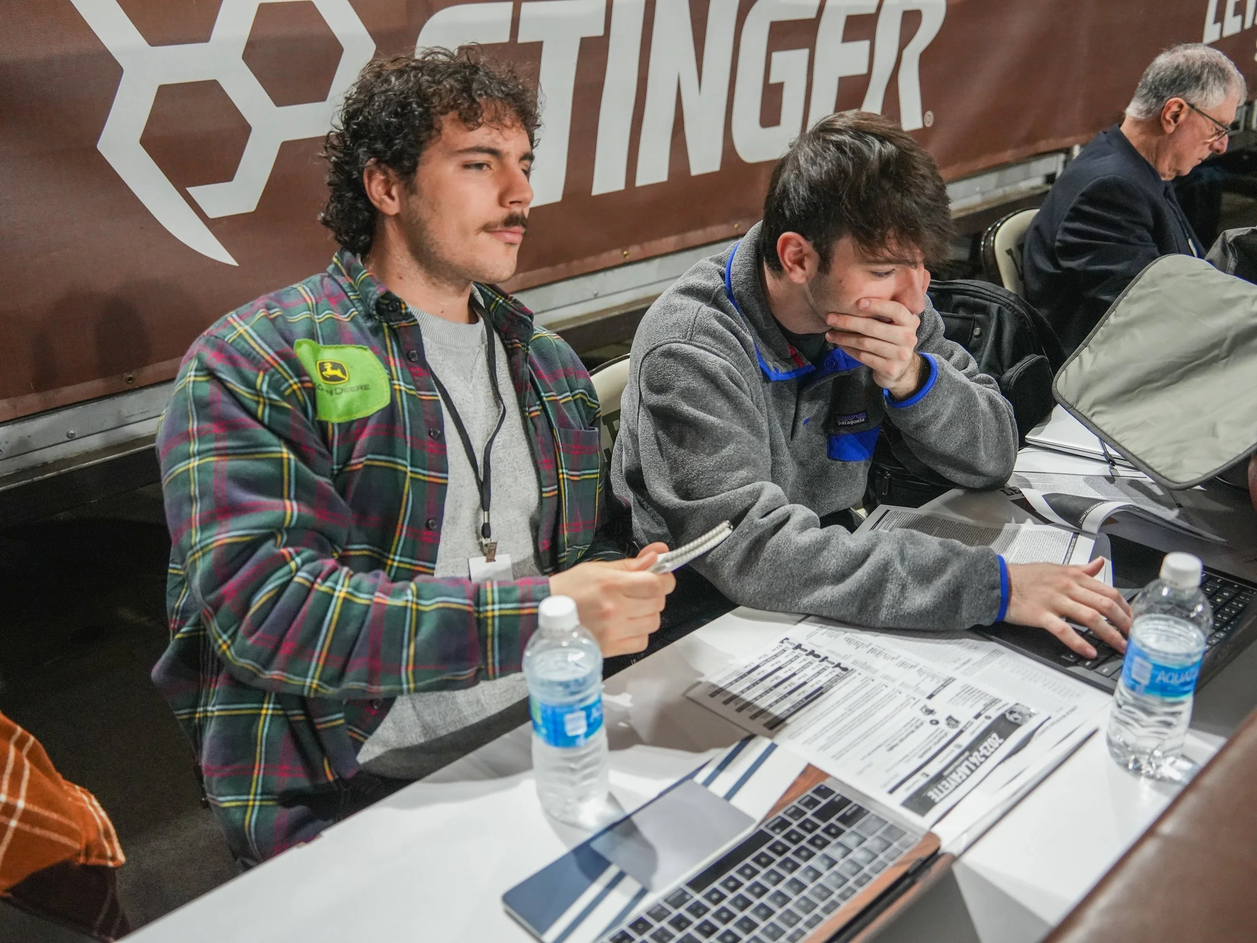 Two men sitting at a table, one with a plaid shirt holding a notebook, the other with a gray fleece covering his hand, looking at their laptops, with water bottles and papers on the table, and a large Rwanda Science logo in the background.