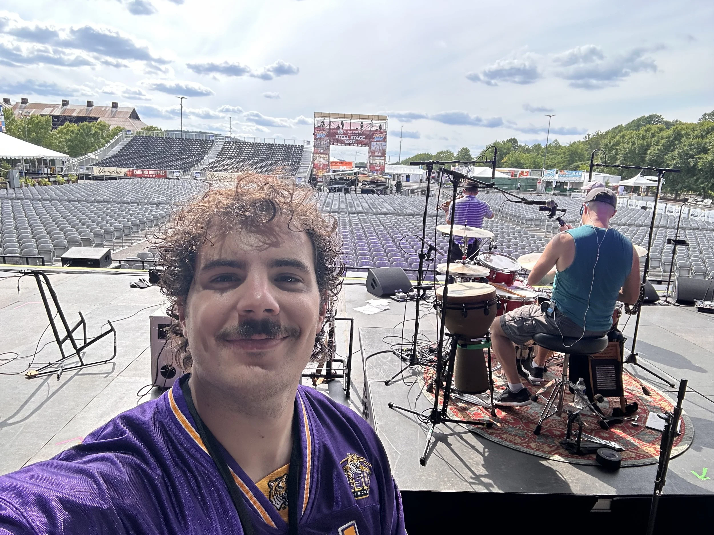 A young man with curly hair and a mustache is taking a selfie on a stage before a large outdoor concert or event. Behind him, a band is warming up, with a drummer sitting at a drum kit. Empty seats and a large screen or banner are visible in the background under partly cloudy skies.