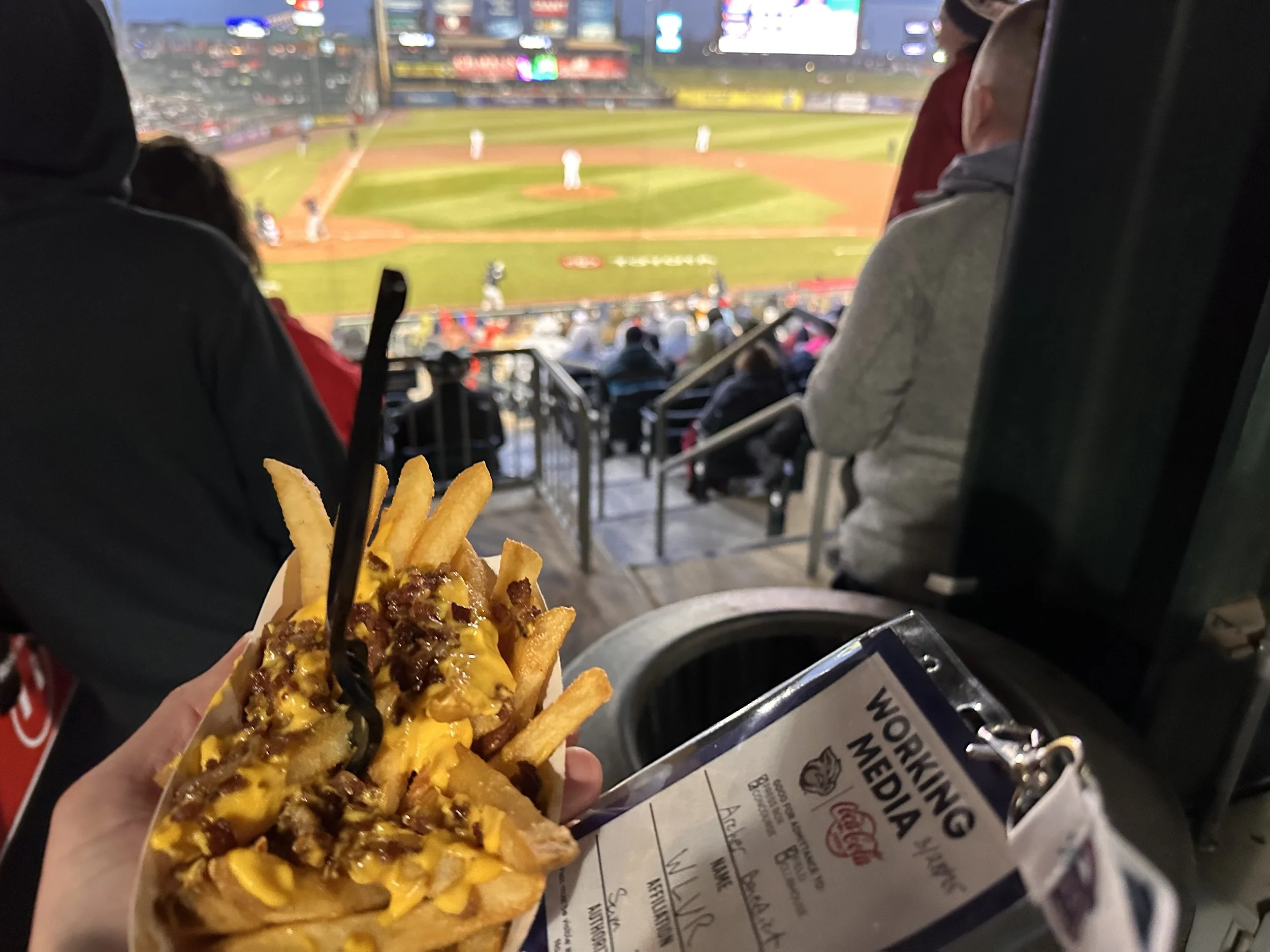 Person holding tray of French fries topped with melted cheese and minced meat at a stadium baseball game with spectators and players in the background.