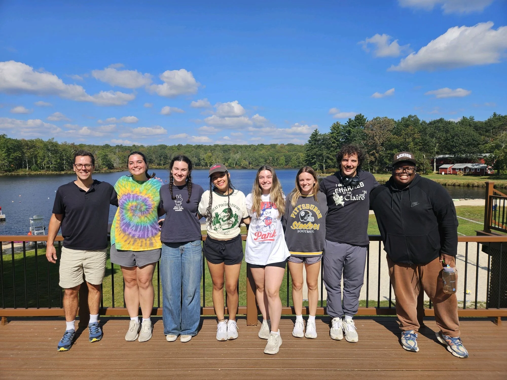 Group of eight young people standing on a wooden deck outdoors, smiling, with a lake, trees, and a blue sky with clouds in the background.