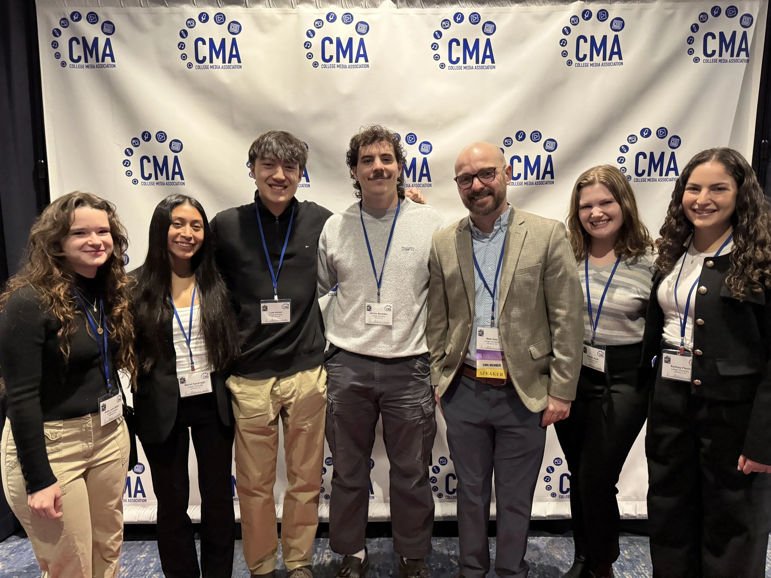 Group of seven young adults and one older man standing together at an event, smiling, with a backdrop displaying the College Media Association (CMA) logo.
