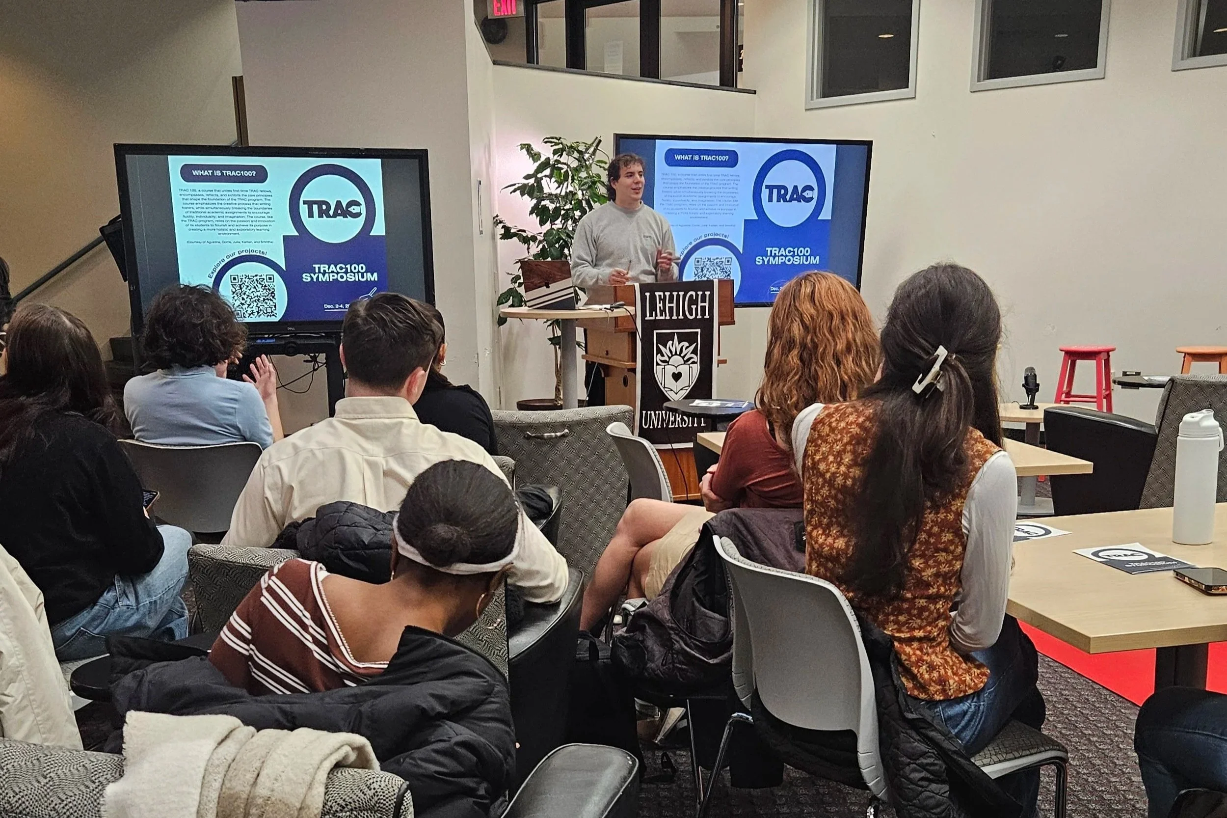 A man giving a presentation at a Trac100 Symposium at Lehigh University, standing behind a podium with a sign reading 'Lehigh University,' in front of two large screens displaying a slide about Trac100.