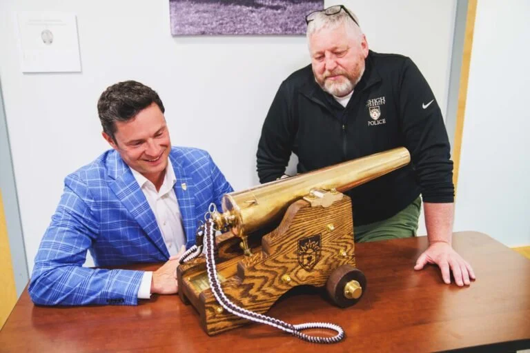 Two men look at a brass cannon replica on a wooden stand, one is smiling and wearing a blue blazer, the other has glasses on head and is wearing a black jacket.