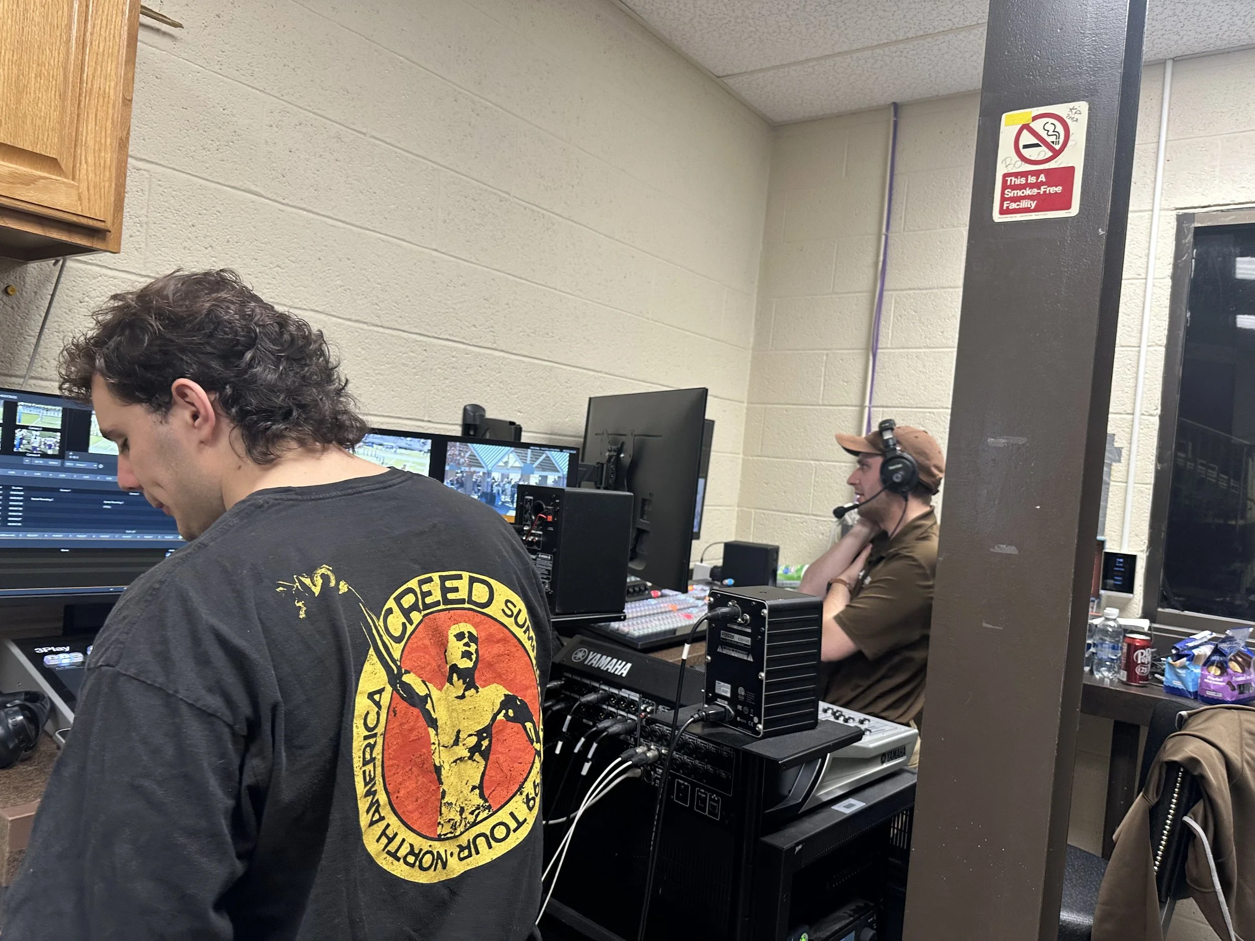 Two men working at a video editing and sound production station in a room. One man with long curly hair wearing a black T-shirt with a graphic of a person on the back. The other man wearing a brown cap and headphones, sitting at a desk with multiple monitors, audio equipment, and supplies.